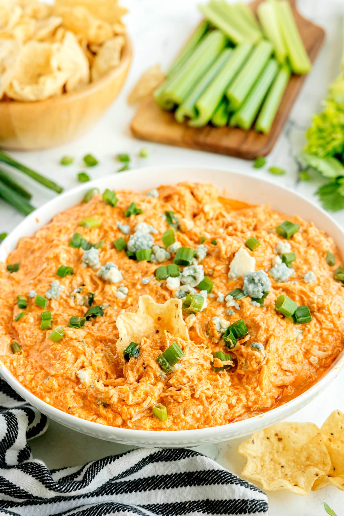 A bowl of High Protein Buffalo Chicken Dip topped with green onions and blue cheese crumbles, surrounded by tortilla chips and celery sticks.