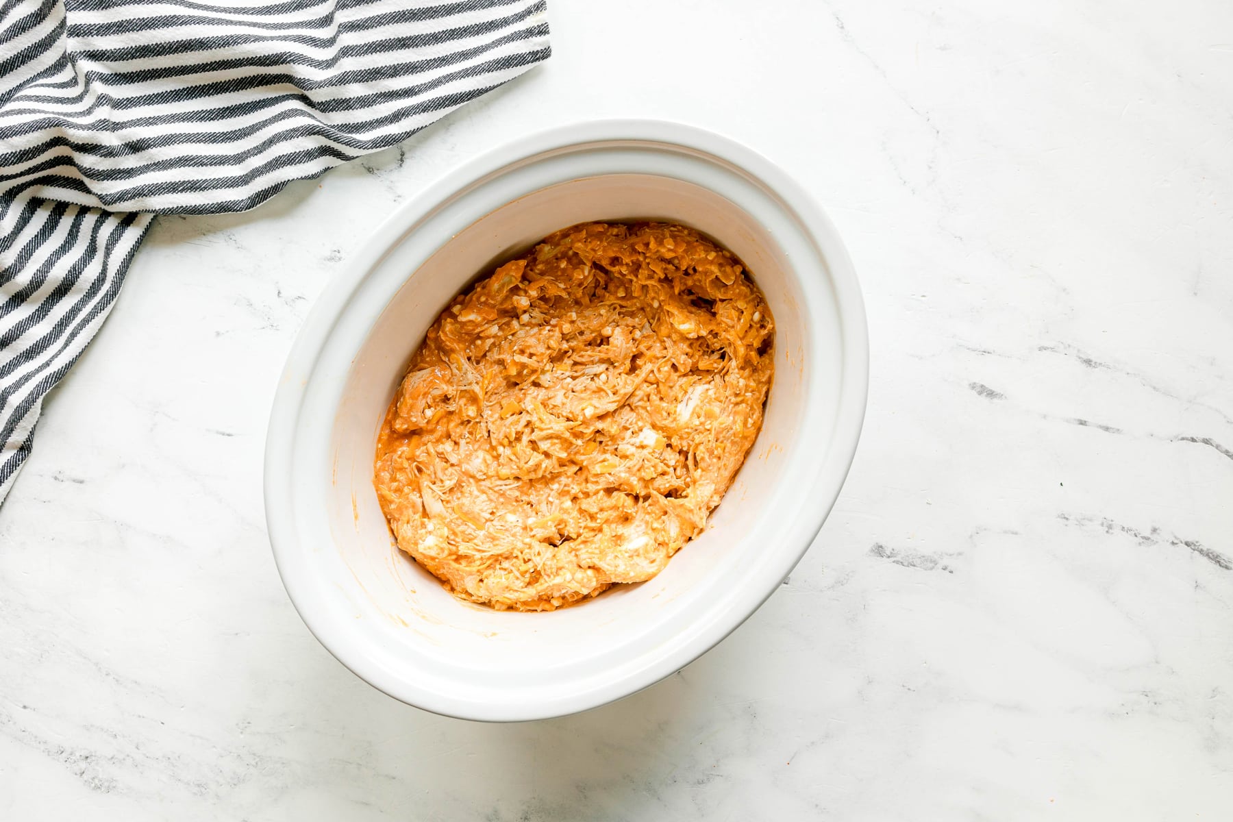 A white slow cooker with High Protein Buffalo Chicken Dip mixture rested on a white marble surface next to a striped cloth.