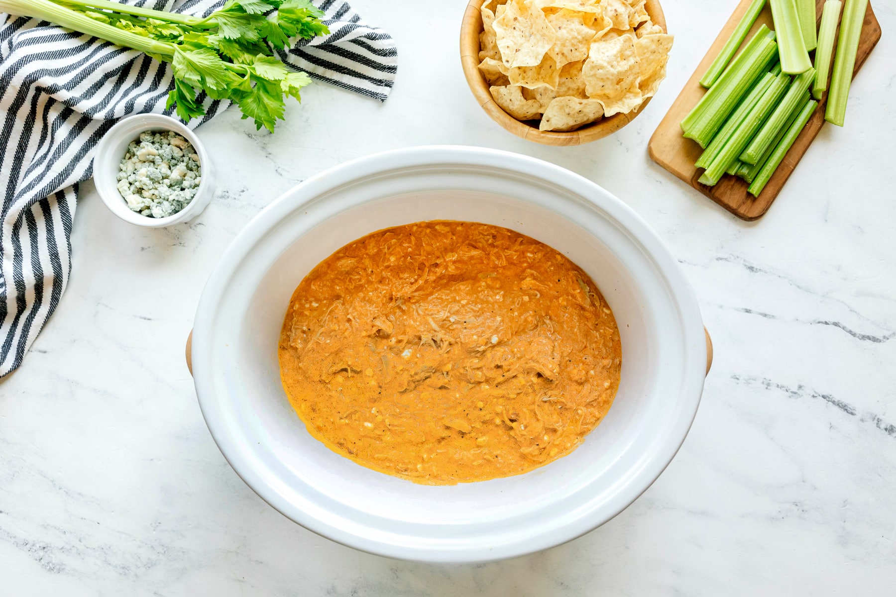 A slow cooker filled with High Protein Buffalo Chicken Dip sits on a marble counter, surrounded by celery sticks, tortilla chips, blue cheese, and a striped towel.