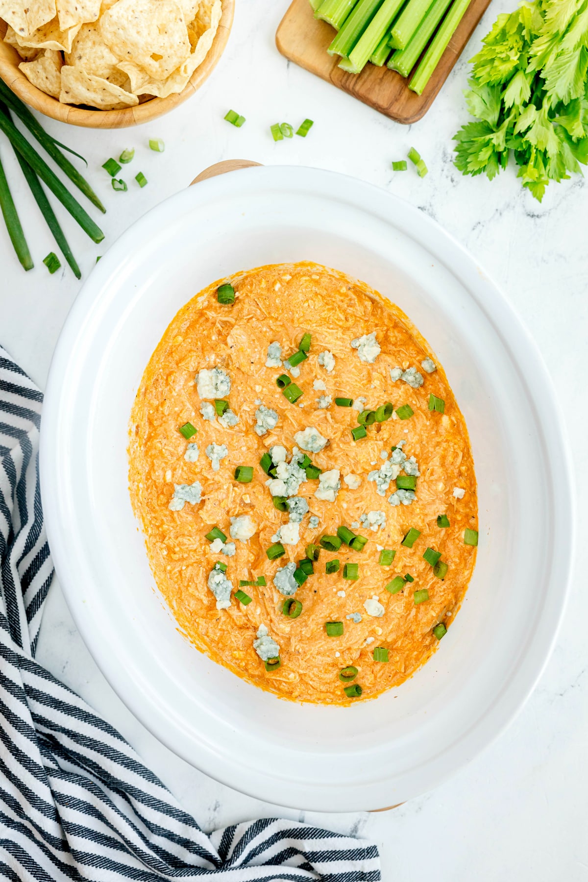 A white oval dish filled with High Protein Buffalo Chicken Dip, topped with chopped green onions and blue cheese, surrounded by celery stalks, tortilla chips, and a striped towel.