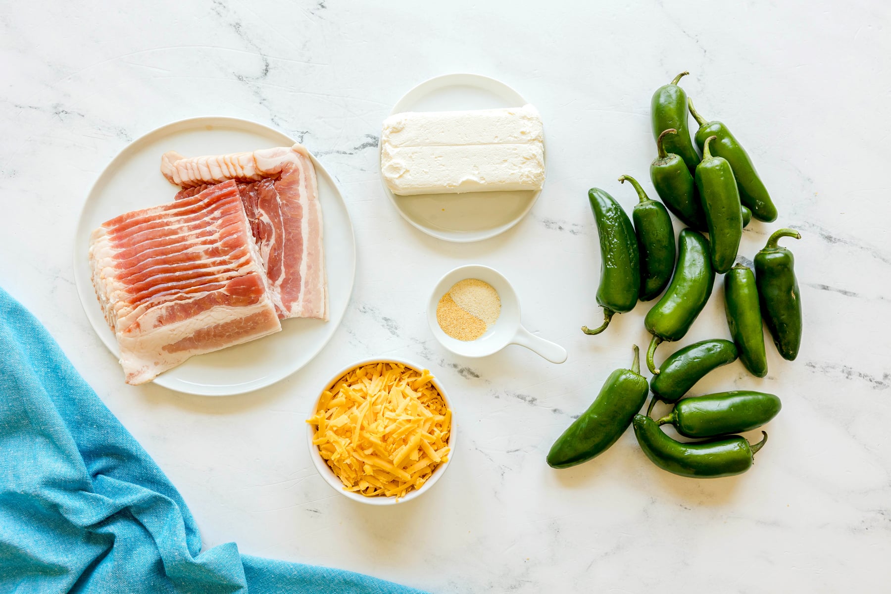 Ingredients for Jalapeño Poppers Wrapped in Bacon are arranged on a white marble surface with a blue cloth. It includes: sliced bacon, shredded cheese, softened cream cheese, fresh jalapeño peppers, and a garlic and onion powder
