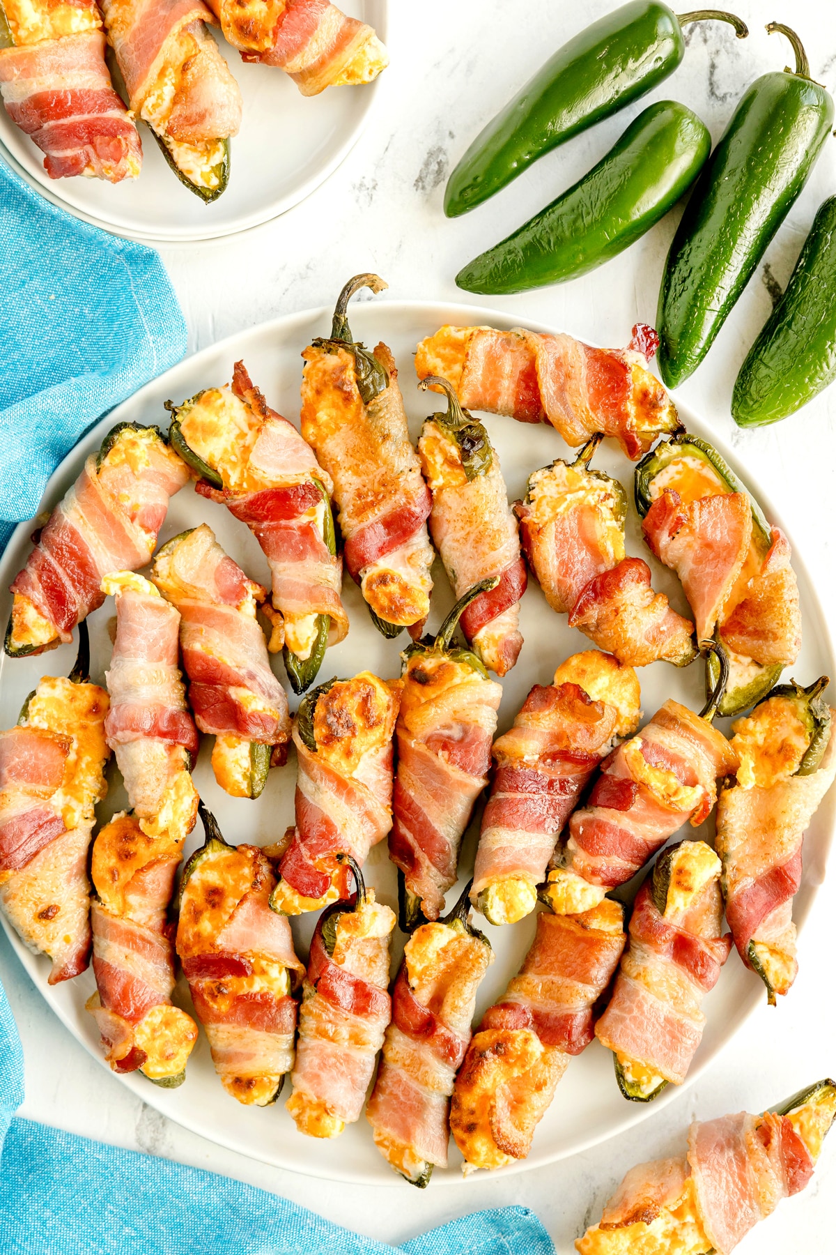 A plate of Jalapeño Poppers Wrapped in Bacon filled with a creamy cheese mixture, with fresh jalapeños and a blue napkin nearby.