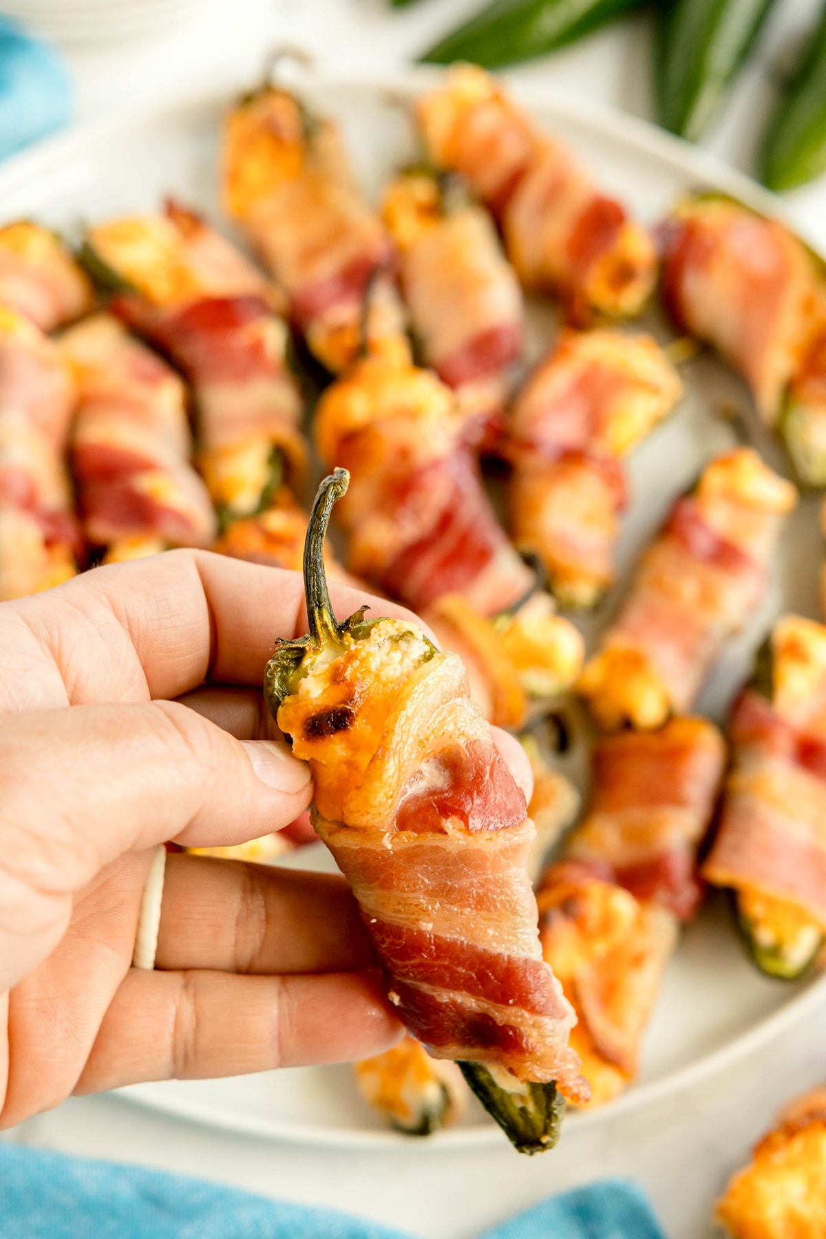 A hand holding a Jalapeño Poppers Wrapped in Bacon filled with cheese; more poppers are arranged on a white plate in the background.
