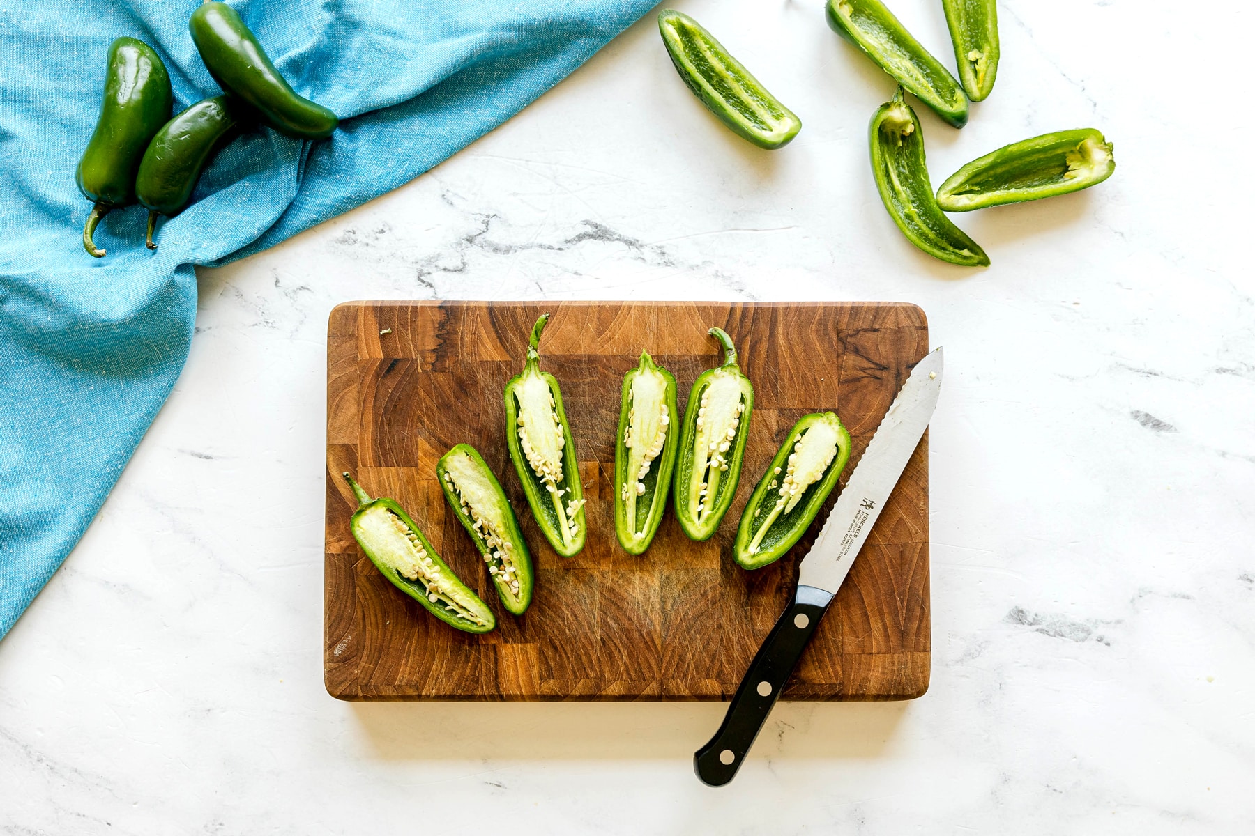 Six halved jalapeños for Jalapeño Poppers Wrapped in Bacon recipe with seeds are on a wooden cutting board next to a knife, with more jalapeños and a blue cloth on a white surface.