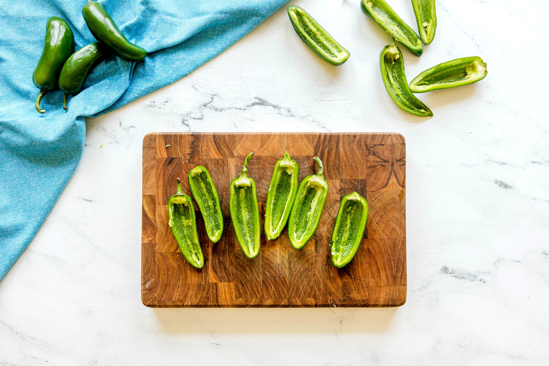 Seeded for Jalapeño Poppers Wrapped in Bacon recipe are arranged on a wooden cutting board, with whole and halved jalapenos and a blue cloth nearby on a marble surface.