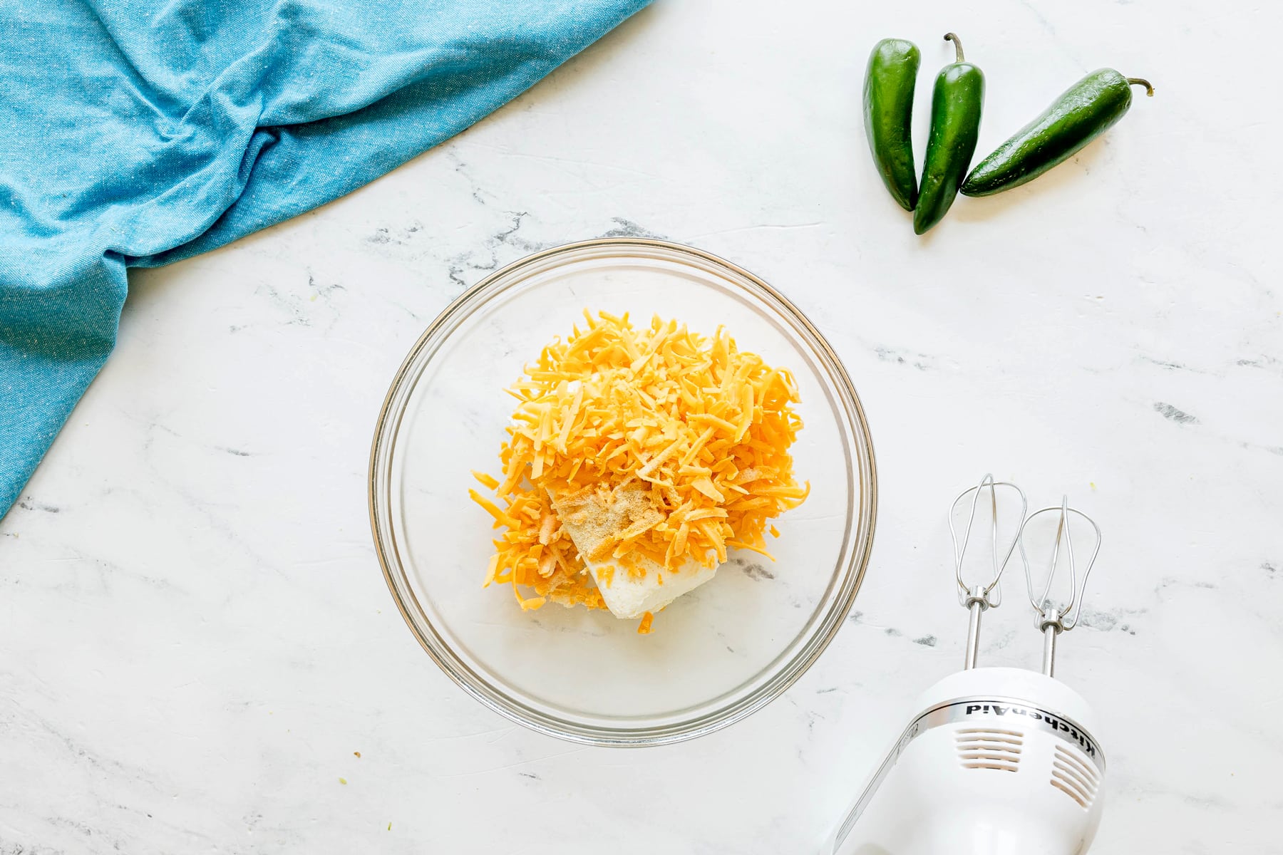 Next step in preparing Jalapeño Poppers Wrapped in Bacon is to use a glass bowl and combine shredded cheese, cream cheese, and garlic and onion powder. It sits on a white surface near two jalapeños, a blue cloth, and a hand mixer.