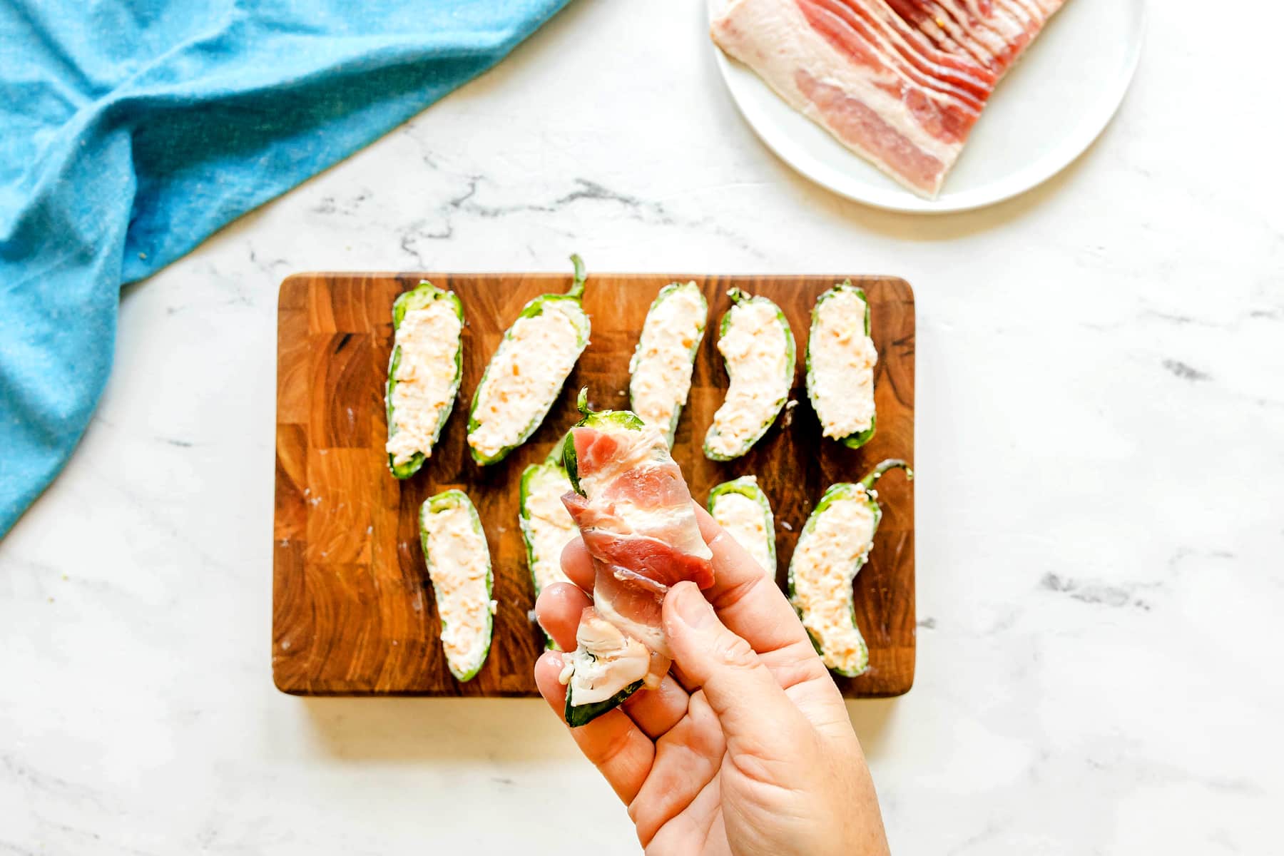 This is the next process in preparing Jalapeño Poppers Wrapped in Bacon. A hand holds a jalapeño half stuffed with filling and wrapped in bacon over a cutting board with more prepared jalapeños; bacon on a plate and blue cloth are in the background.