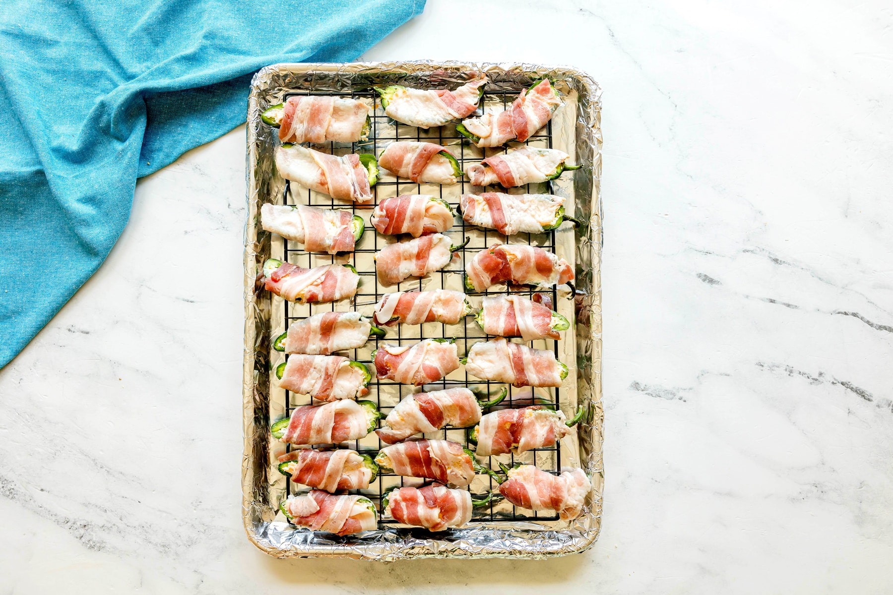 A baking tray lined with foil holds rows of Jalapeño Poppers Wrapped in Bacon on a wire rack, next to a blue cloth on a marble surface.