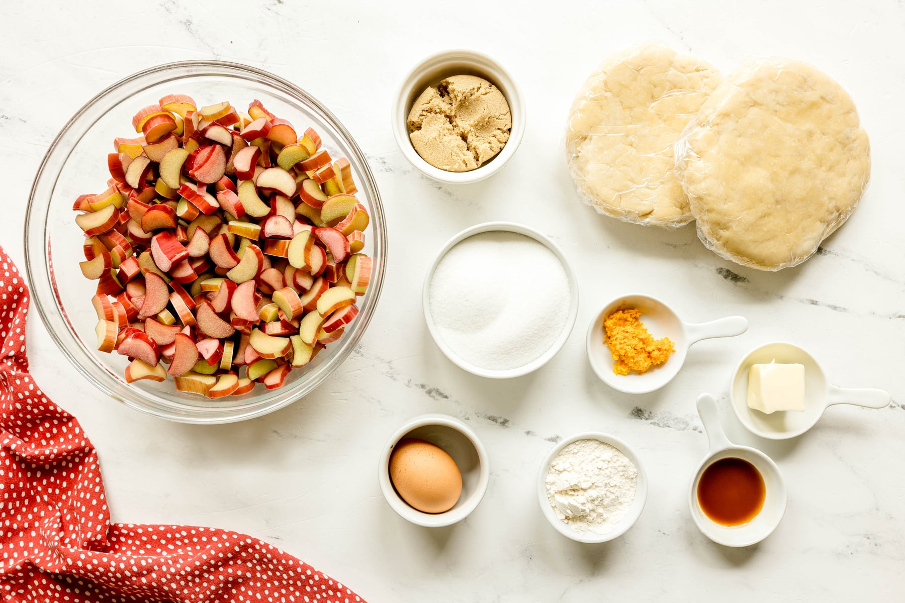 Overhead view of pie ingredients on a white counter, including chopped rhubarb, brown and white sugar, pie dough, egg, flour, orange zest, butter, and vanilla extract.