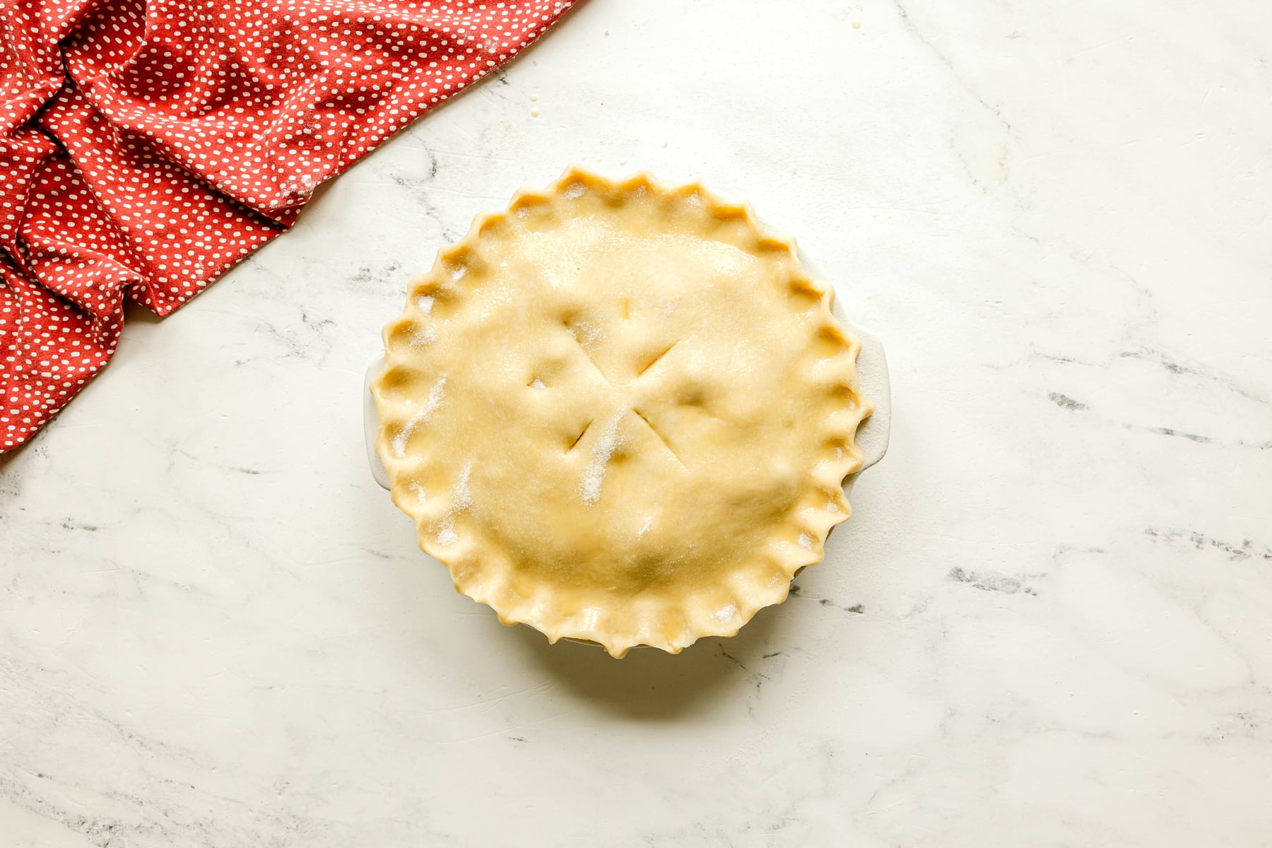 Unbaked Rhubarb Pie with a crimped edge and vent slits sits on a marble surface next to a red polka dot cloth.