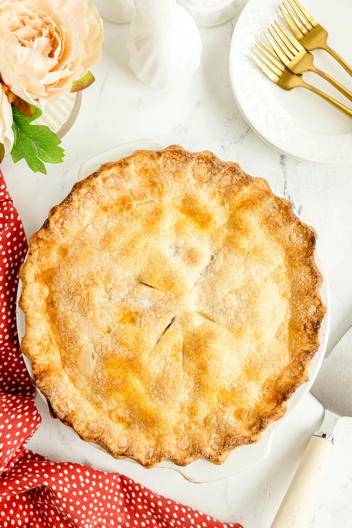 A baked pie with a golden-brown crust sits on a white surface next to a red polka dot napkin, gold forks, white plates, and a flower arrangement.