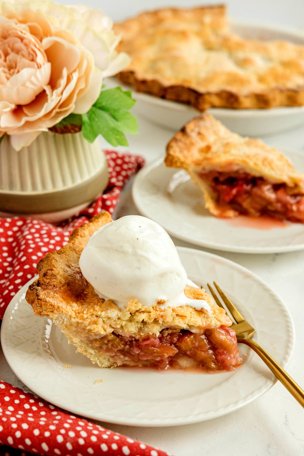 A slice of fruit pie topped with a scoop of vanilla ice cream is served on a plate with a fork, with a whole pie and a flower arrangement in the background.