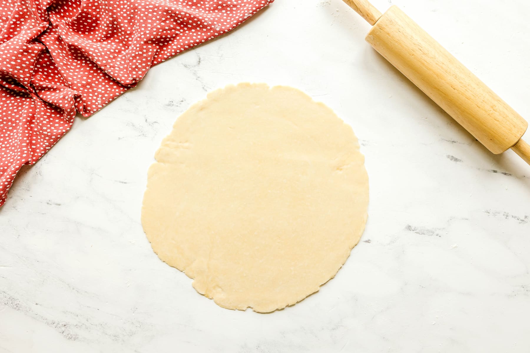 Also another step in preparing Rhubarb Pie Recipe is to roll out dough on a white surface. A wooden rolling pin and a folded red polka dot cloth can also be seen.