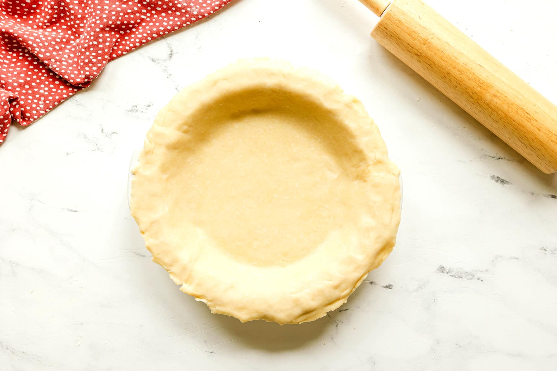 Unbaked pie crust for Rhubarb Pie Recipe on a round dish on a white surface, with a rolling pin and a red polka dot cloth nearby.