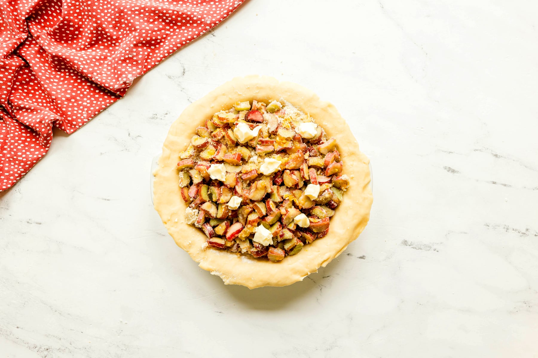 Unbaked Rhubarb Pie filled with chopped fruit and dotted with butter, sitting on a light marble surface next to a red polka dot cloth.