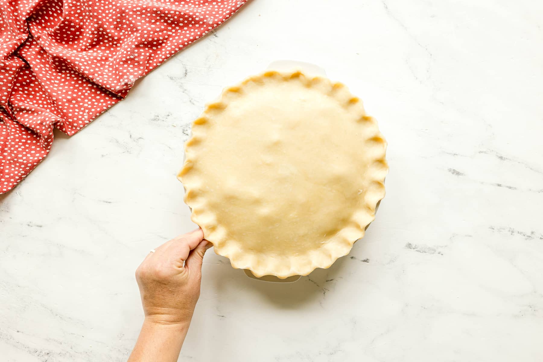 A hand holds an unbaked Rhubarb Pie with a crimped edge on a marble surface, next to a red and white polka dot cloth.