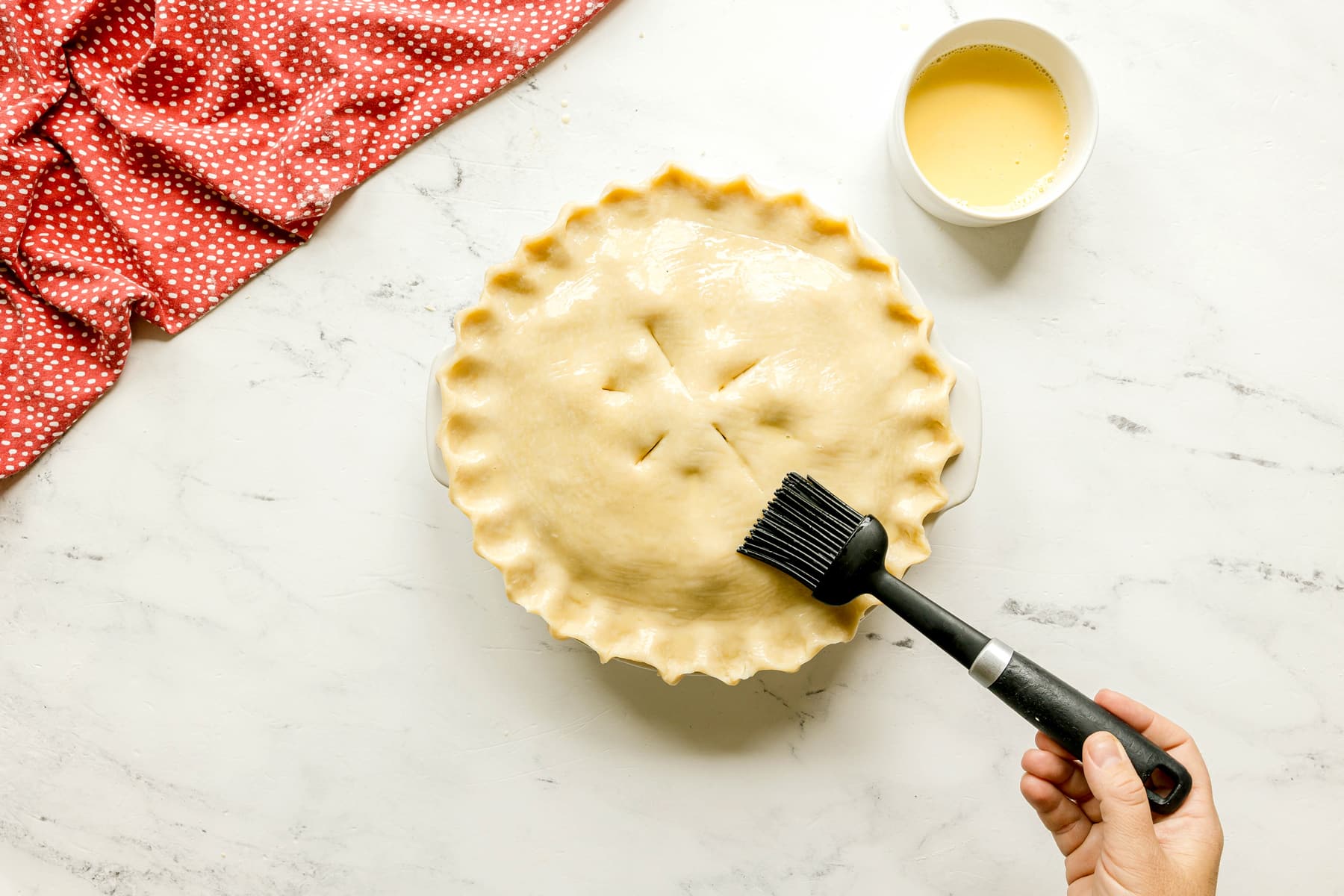 A hand brushes egg wash onto an unbaked Rhubarb Pie crust in a pie dish, with a bowl of egg wash and a red cloth nearby on a marble surface.
