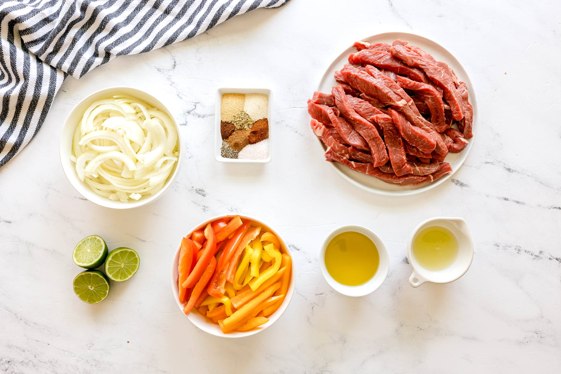Ingredients for Sheet Pan Steak Fajitas are skirt steak, bell peppers, onions, olive oil, onion powder, garlic powder, cumin, chilli powder, chipotle powder, dried oregano, salt and black pepper and lime juice are laid out on a marble surface.