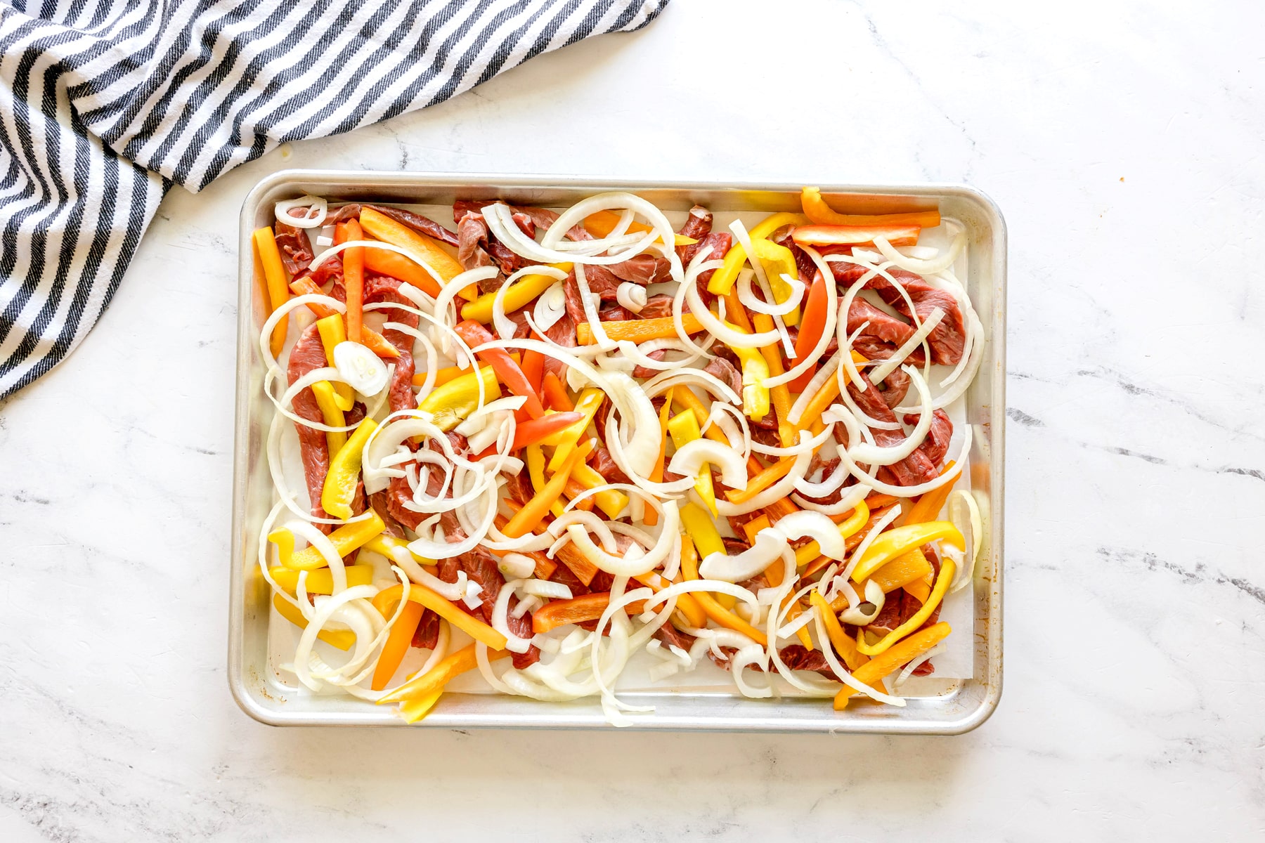 First step in preparing Sheet Pan Steak Fajitas is to add sliced beef, bell peppers, and onions spread on a sheet pan, ready for baking.