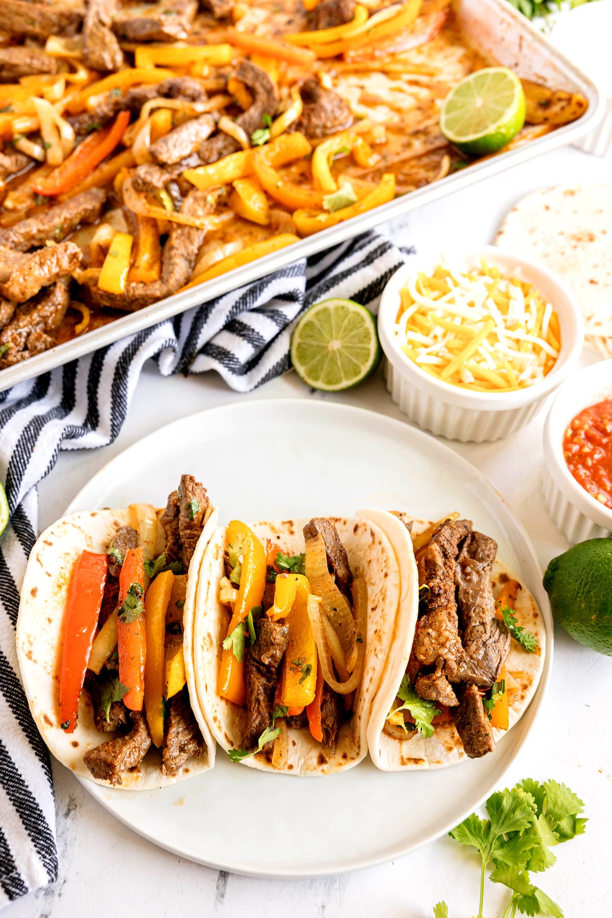 Three beef fajita tacos with bell peppers and onions on a plate, next to lime halves, shredded cheese, salsa, and a sheet pan of flavorful sheet pan steak fajitas.