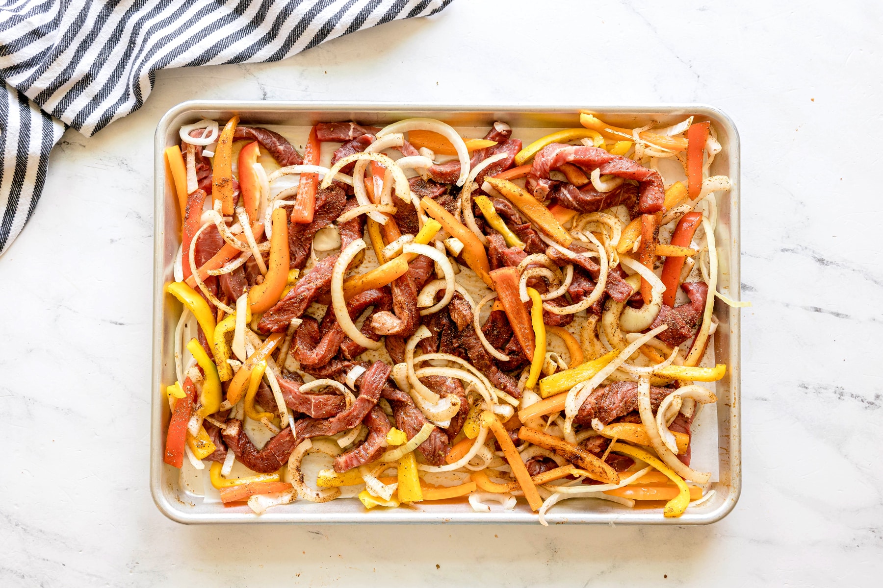 The next step in Sheet Pan Steak Fajitas recipe is to add an olive oil on sheet pan with raw beef strips, sliced onions, and bell peppers, seasoned with spices. Sprinkle this evenly into the pan.