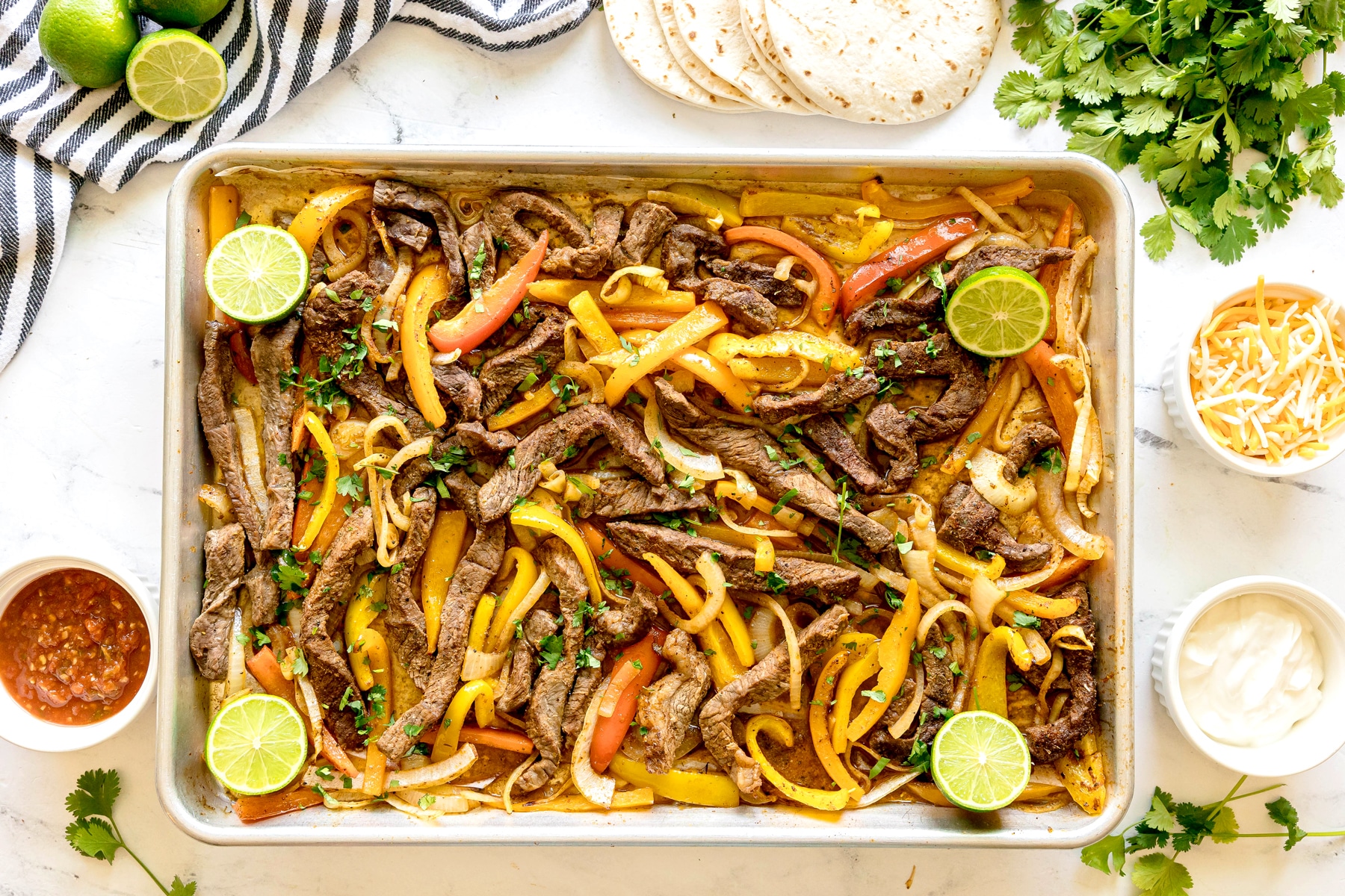 Sheet Pan Steak Fajitas with limes, cilantro, tortillas, salsa, and cheese.