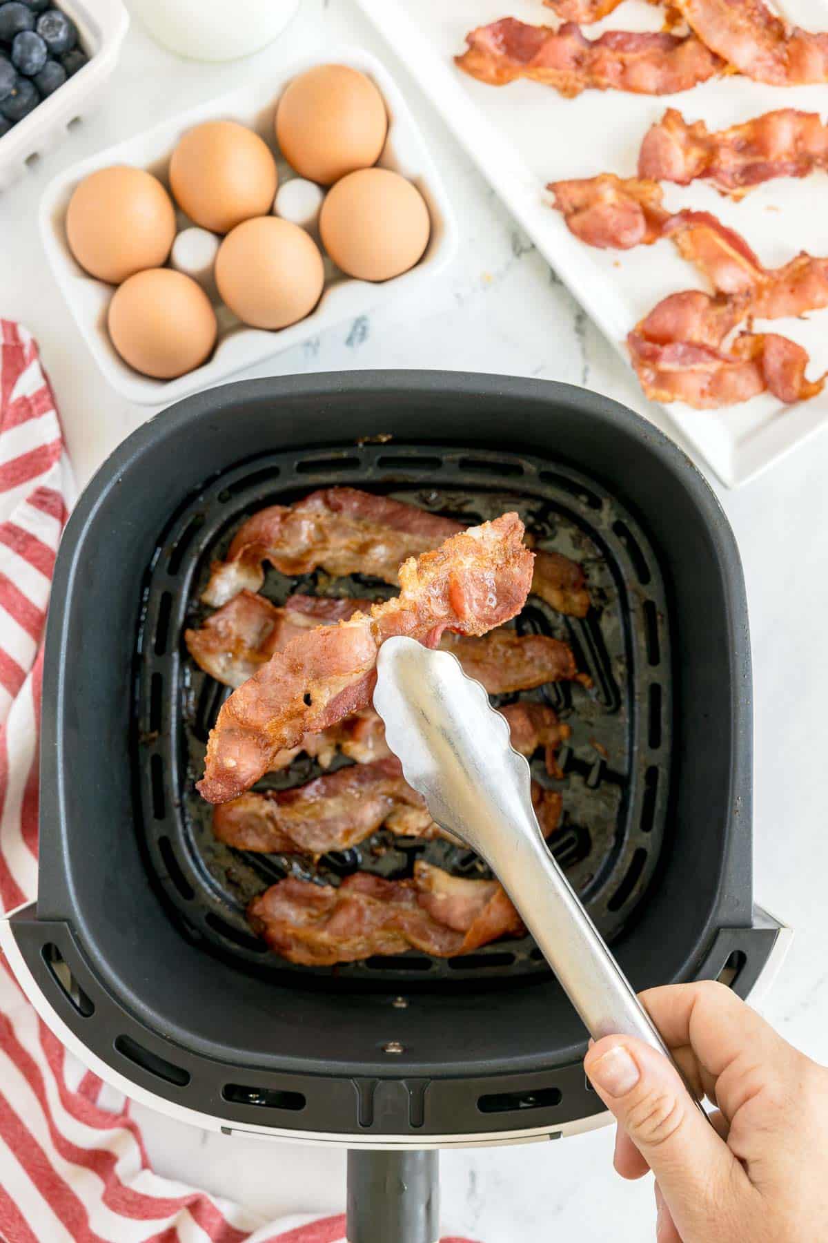 A hand uses tongs to remove cooked bacon from an air fryer basket, with eggs, bacon strips, blueberries, and a red striped towel nearby on the counter.