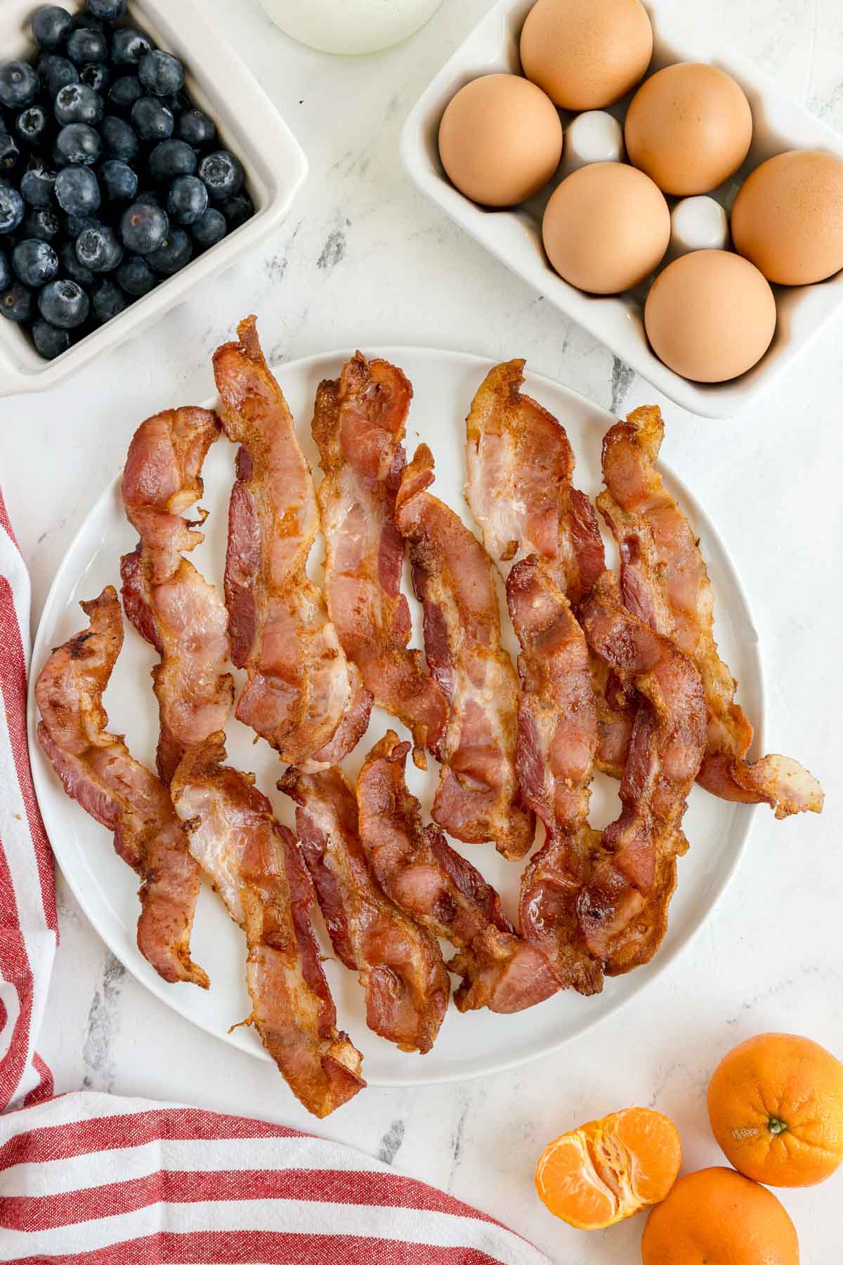 A plate of cooked bacon strips is on a marble surface next to a bowl of eggs, a bowl of blueberries, and two whole tangerines with one peeled segment.