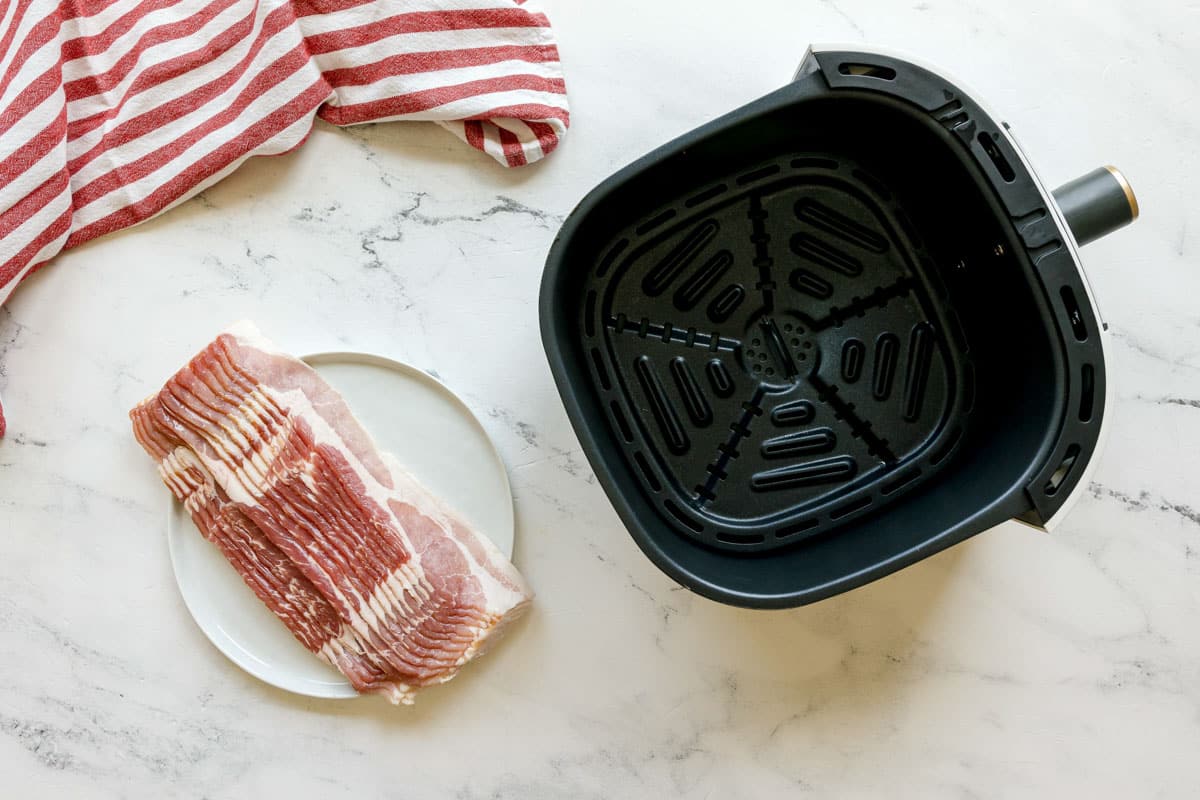 Uncooked bacon on a white plate next to an empty air fryer basket on a marble countertop with a red and white striped towel.