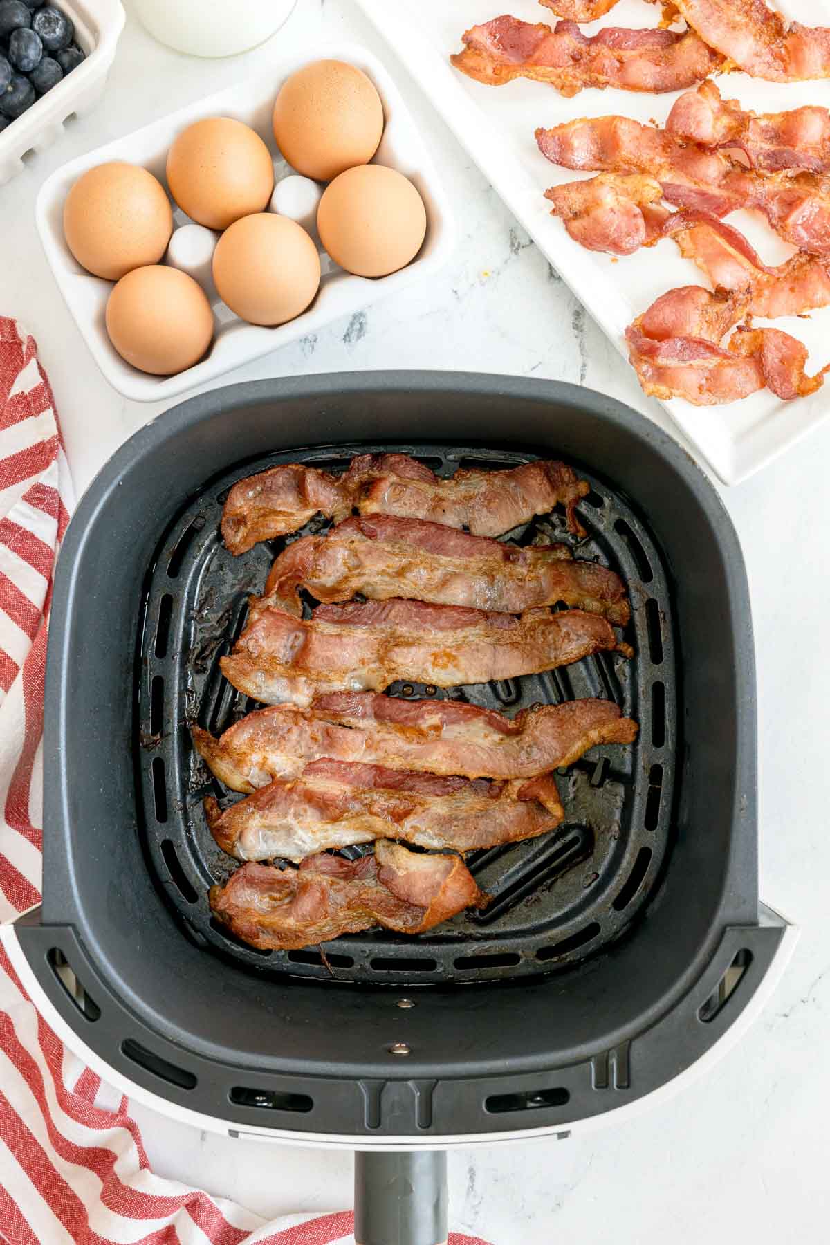 Cooked bacon strips in an air fryer basket, with raw eggs, cooked bacon on a plate, blueberries, and a red-striped towel nearby on a white surface.