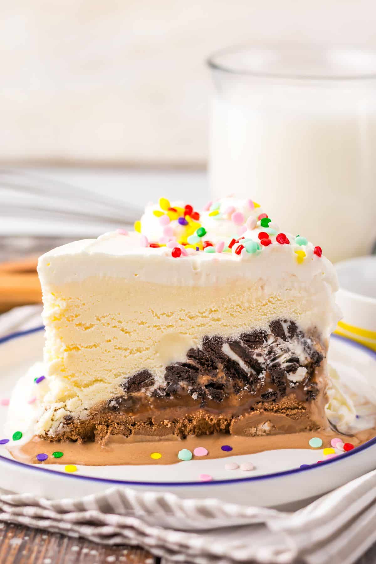 A slice of layered ice cream cake with chocolate and vanilla, topped with whipped cream and colorful sprinkles, on a plate with a glass of milk in the background.