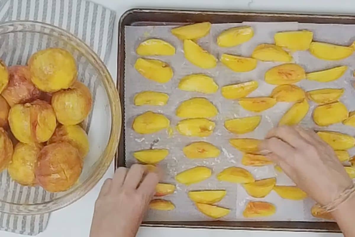 Hands arrange sliced yellow peaches on a parchment-lined baking sheet; whole peeled peaches are in a glass bowl nearby on a striped cloth.
