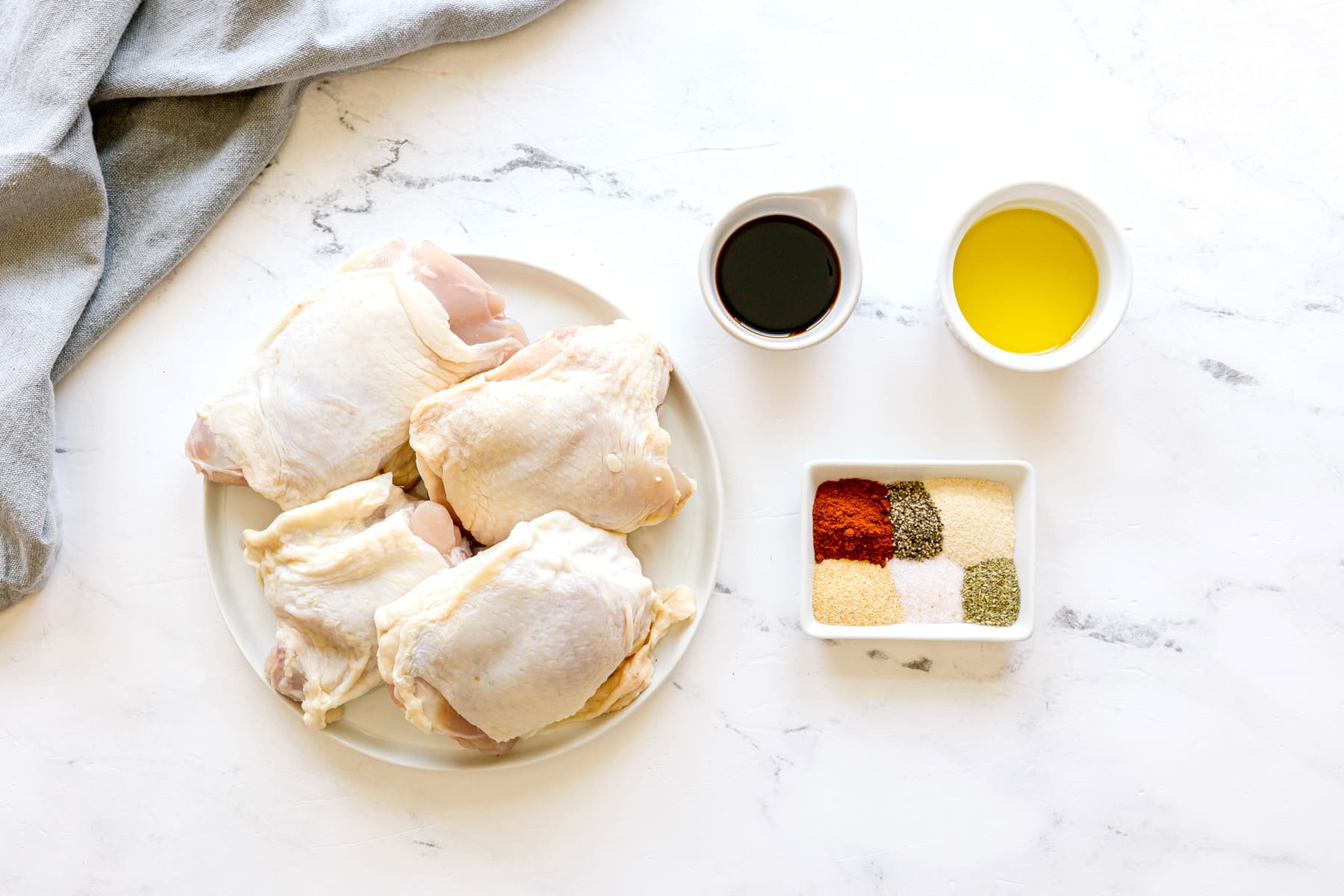 Ingredients for Air Fryer Chicken Thighs: raw chicken thighs on a plate, with small bowls containing olive oil, soy sauce, paprika, garlic powder, kosher salt, black pepper, and Italian seasoning on a white marble surface.