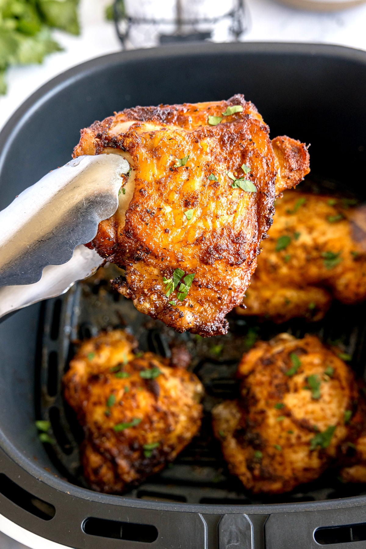 A pair of tongs holds a seasoned, cooked Air Fryer Chicken Thighs above other pieces inside an air fryer basket.