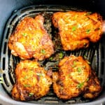 Four cooked, seasoned Air Fryer Chicken Thighs sit in an air fryer basket, garnished with chopped herbs.