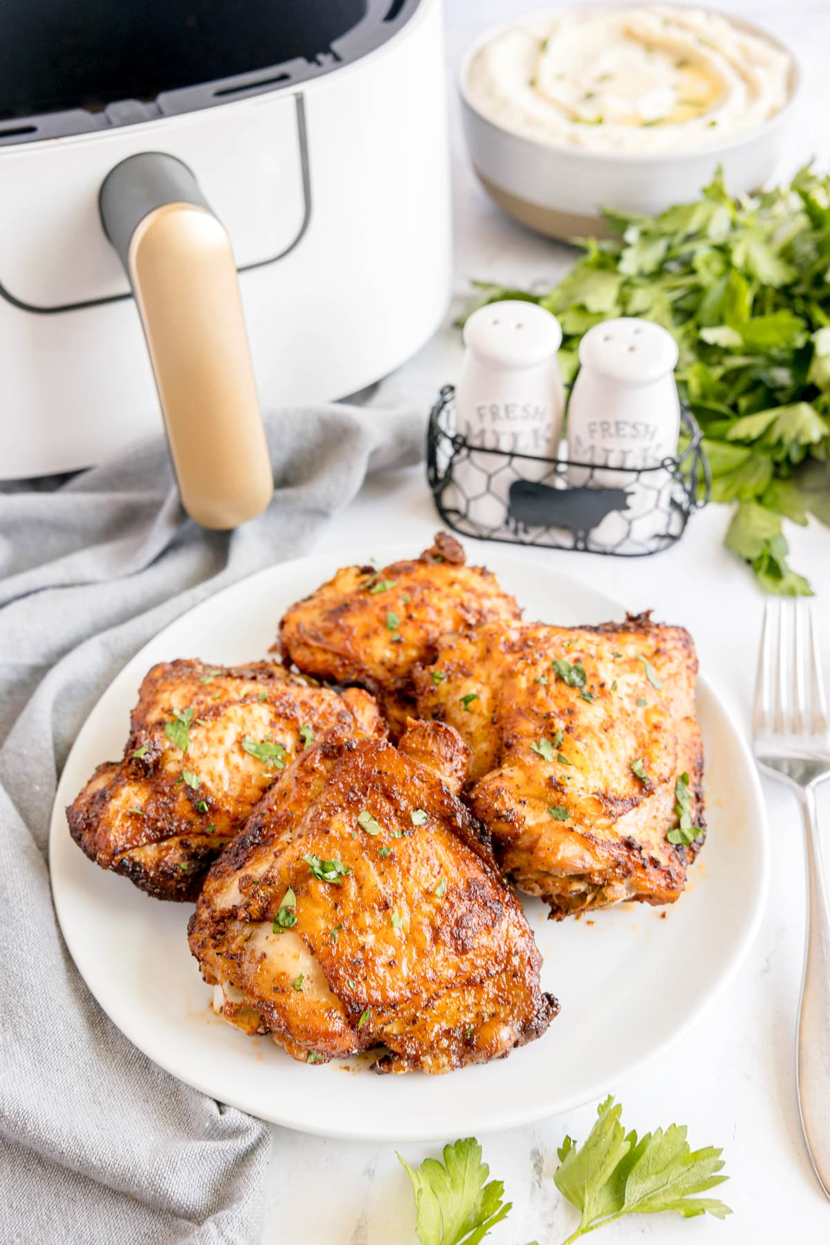 Four pieces of seasoned, cooked Air Fryer Chicken Thighs on a white plate, next to an air fryer, salt and pepper shakers, mashed potatoes, and fresh parsley.