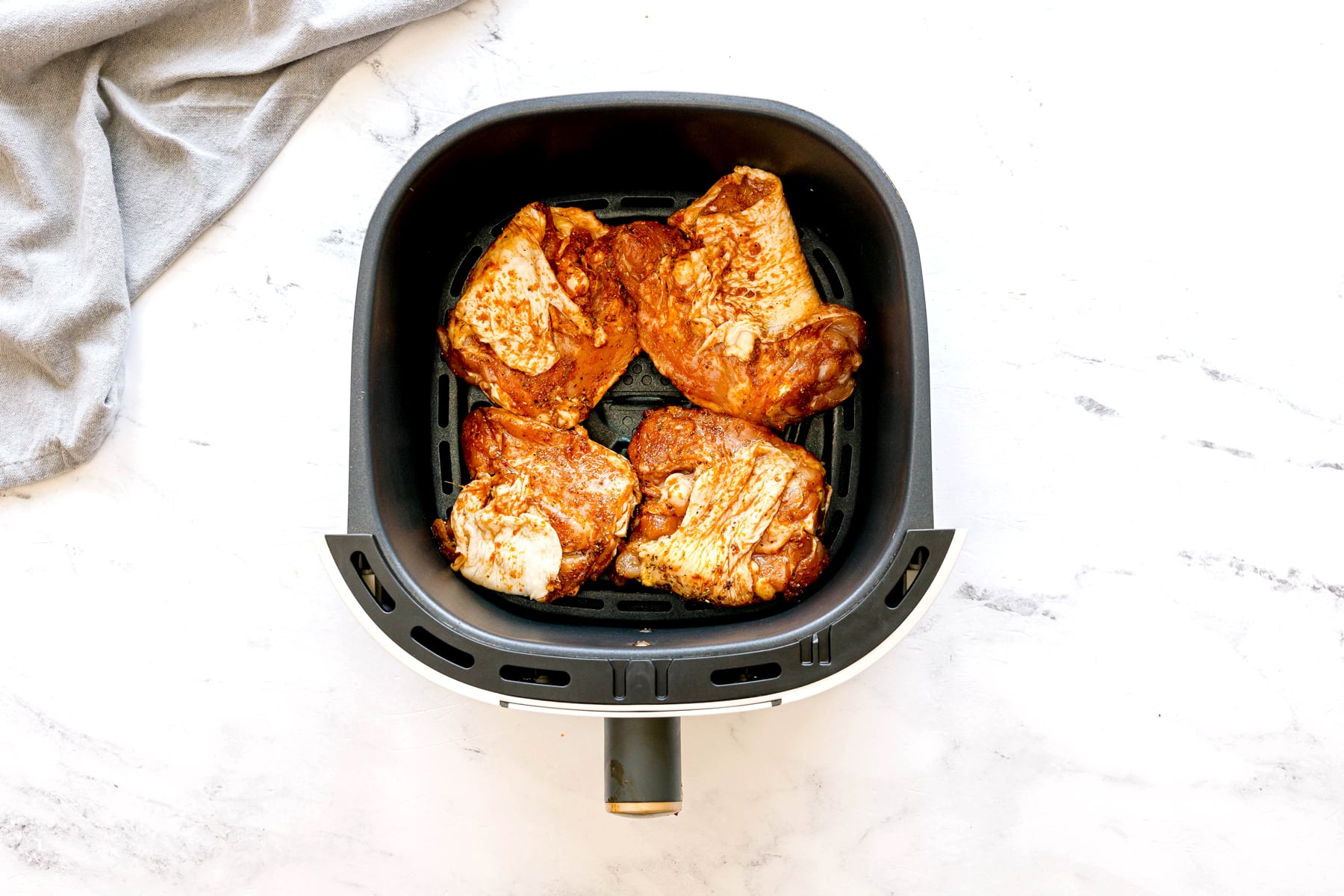 Four seasoned chicken thighs are arranged inside an air fryer basket on a white marble surface, with a gray cloth in the upper left corner. This is one of the steps in preparing Air Fryer Chicken Thighs