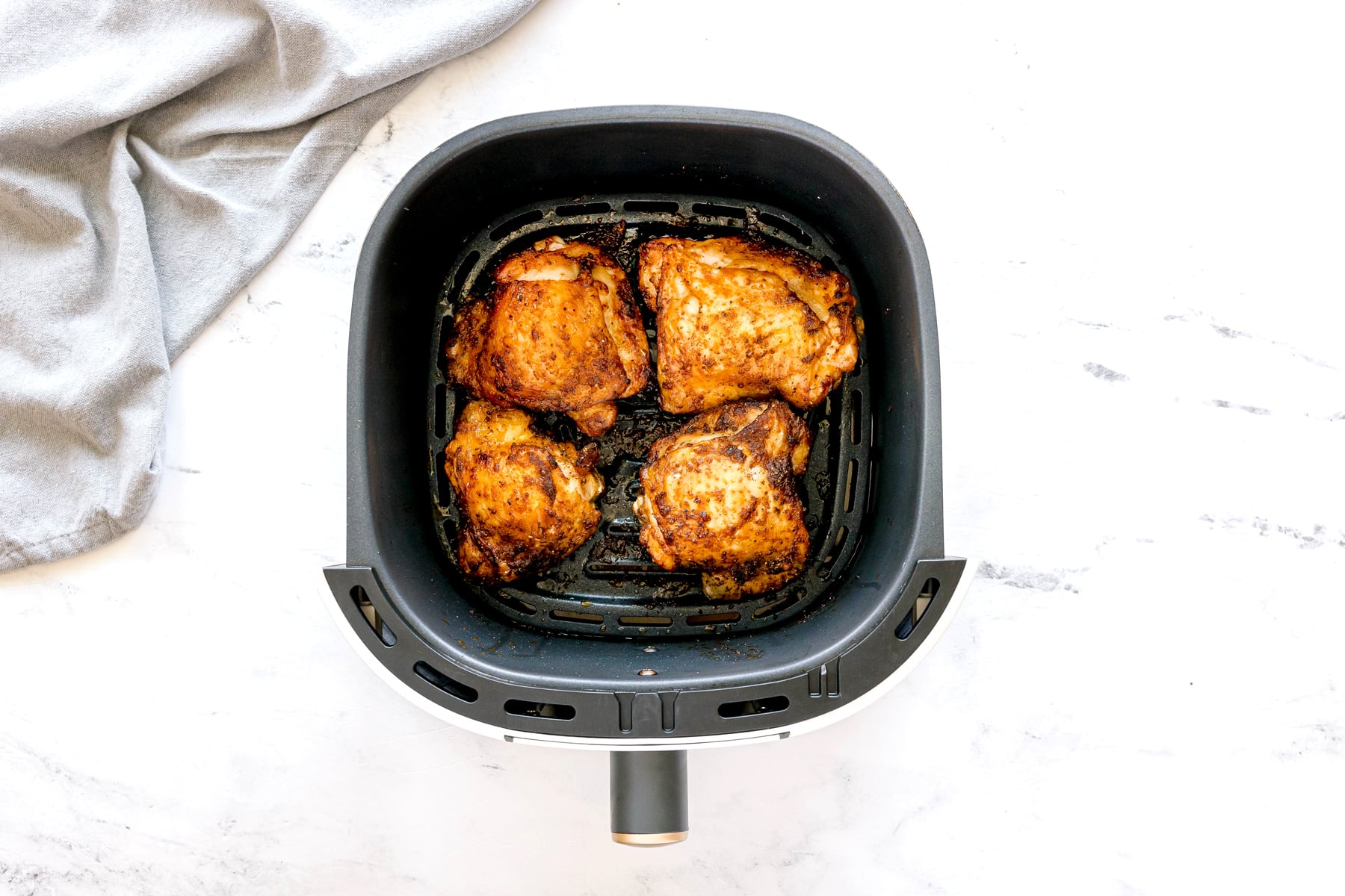 Four Air Fryer Chicken Thighs in an air fryer basket, viewed from above, on a white countertop with a gray cloth nearby.