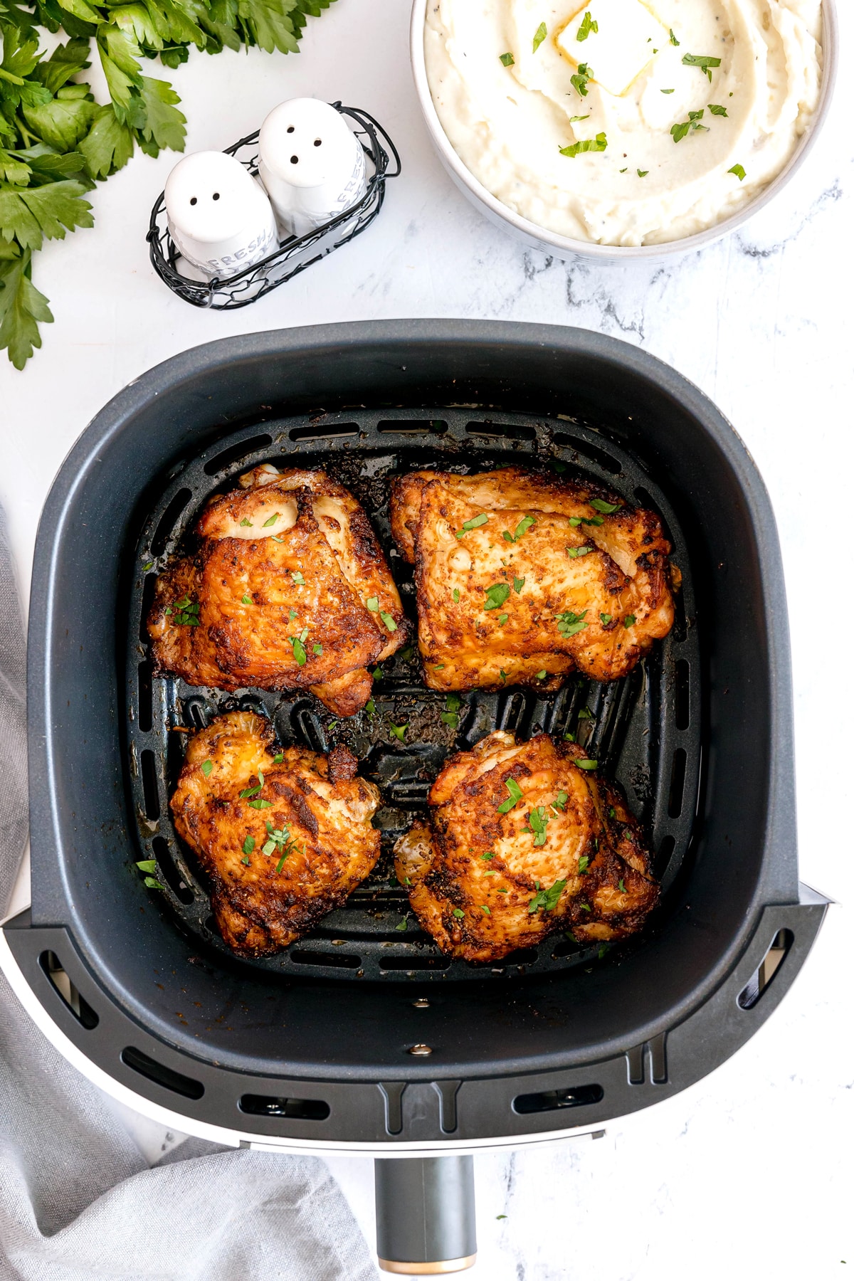 Four cooked Air Fryer Chicken Thighs with seasoning in an air fryer basket, with mashed potatoes and parsley in the background.