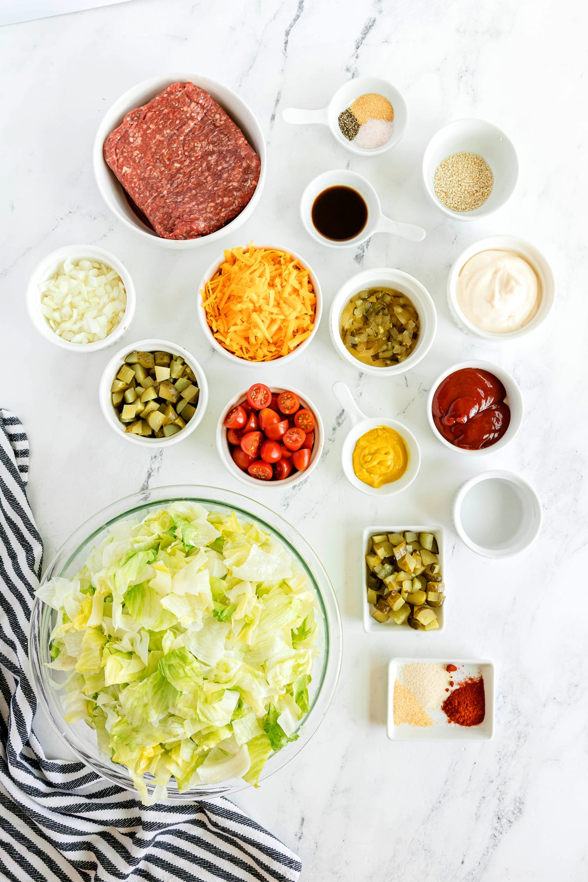 Ingredients for Big Mac Salad includes a glass bowl with mayonnaise, ketchup, yellow mustard, sweet pickle relish, dill pickles, white vinegar, paprika, onion powder, and garlic powder iceberg lettuce, Worcestershire sauce, shredded cheddar cheese, dill pickles,cherry tomatoes, sesame seeds, sits on a white surface next to a bowl of cooked ground beef and a striped cloth.
