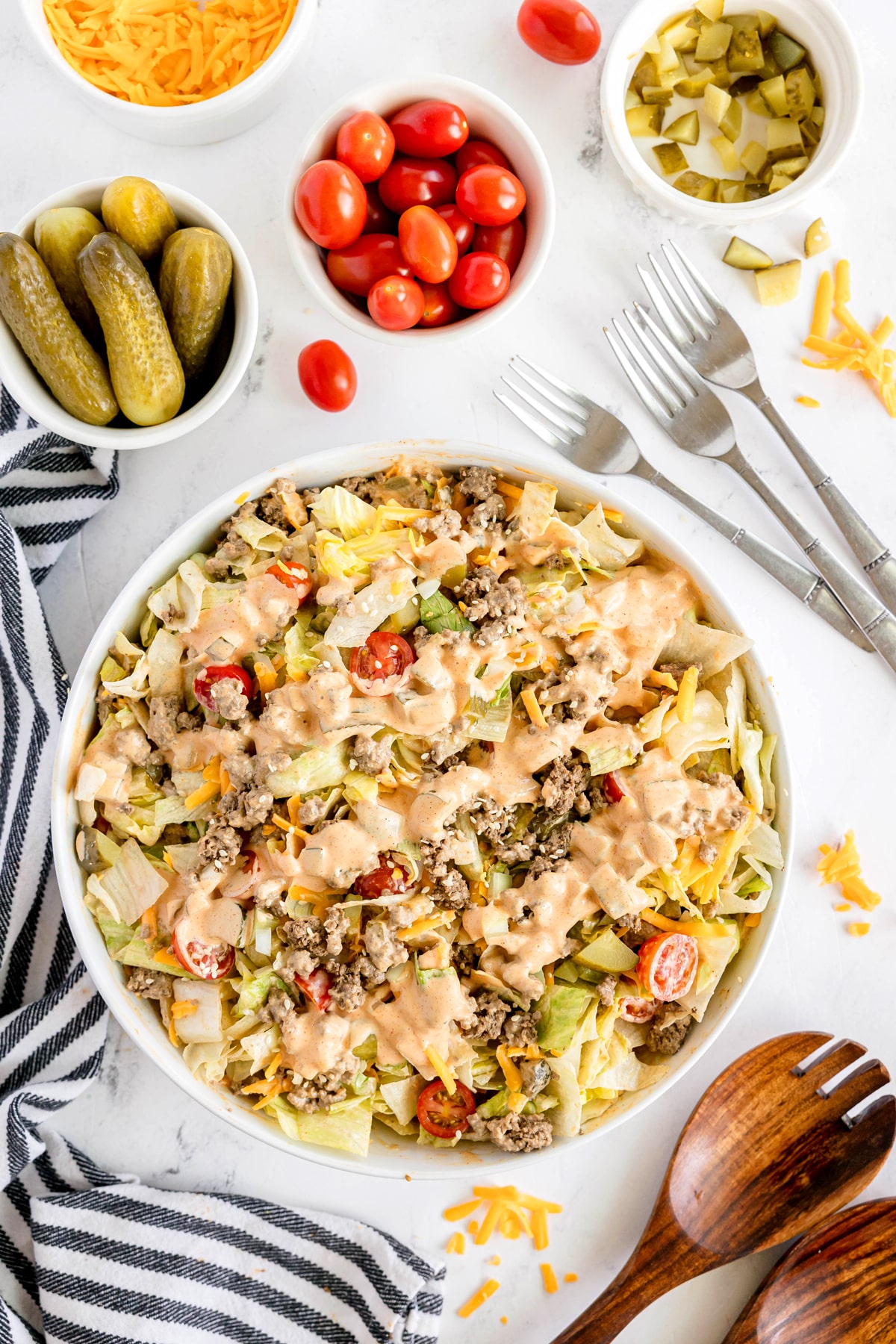 A bowl of Big Mac Salad with ground beef, lettuce, tomatoes, shredded cheese, pickles, and dressing, surrounded by small bowls of cherry tomatoes, pickles, cheese, and sliced pickles.
