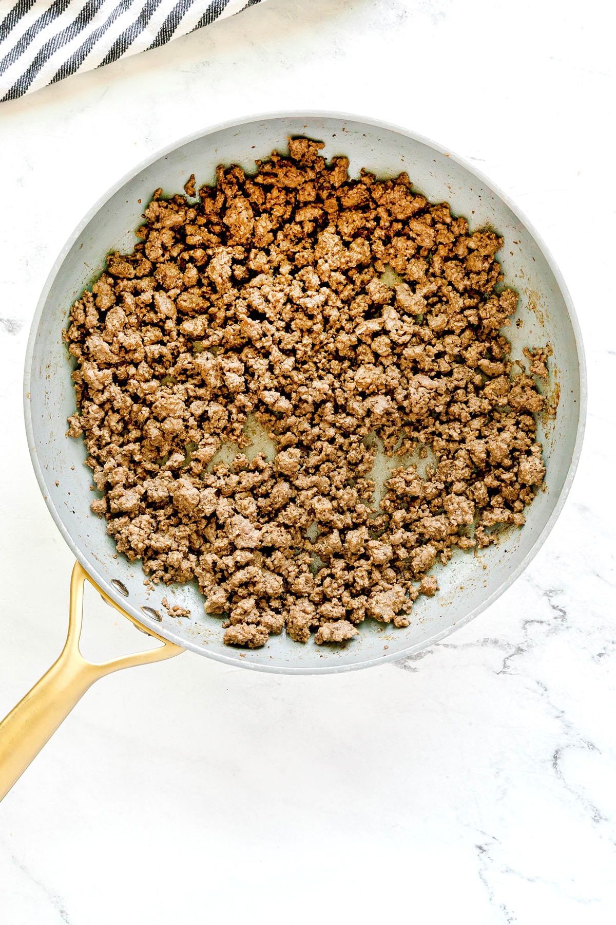 Cooked ground beef in a frying pan on a white marble surface, perfect for a Big Mac Salad Recipe, with a striped towel partially visible at the top left corner.