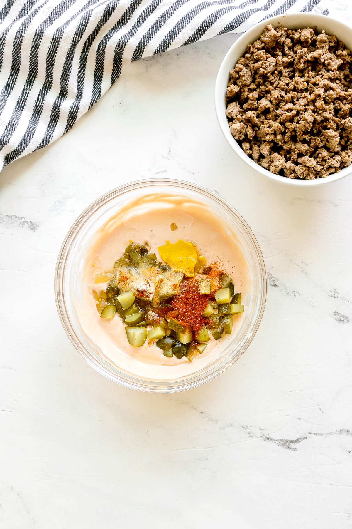 A glass bowl with mayonnaise, chopped pickles, mustard, and spices sits on a white surface next to a bowl of cooked ground beef for Big Mac Salad recipe