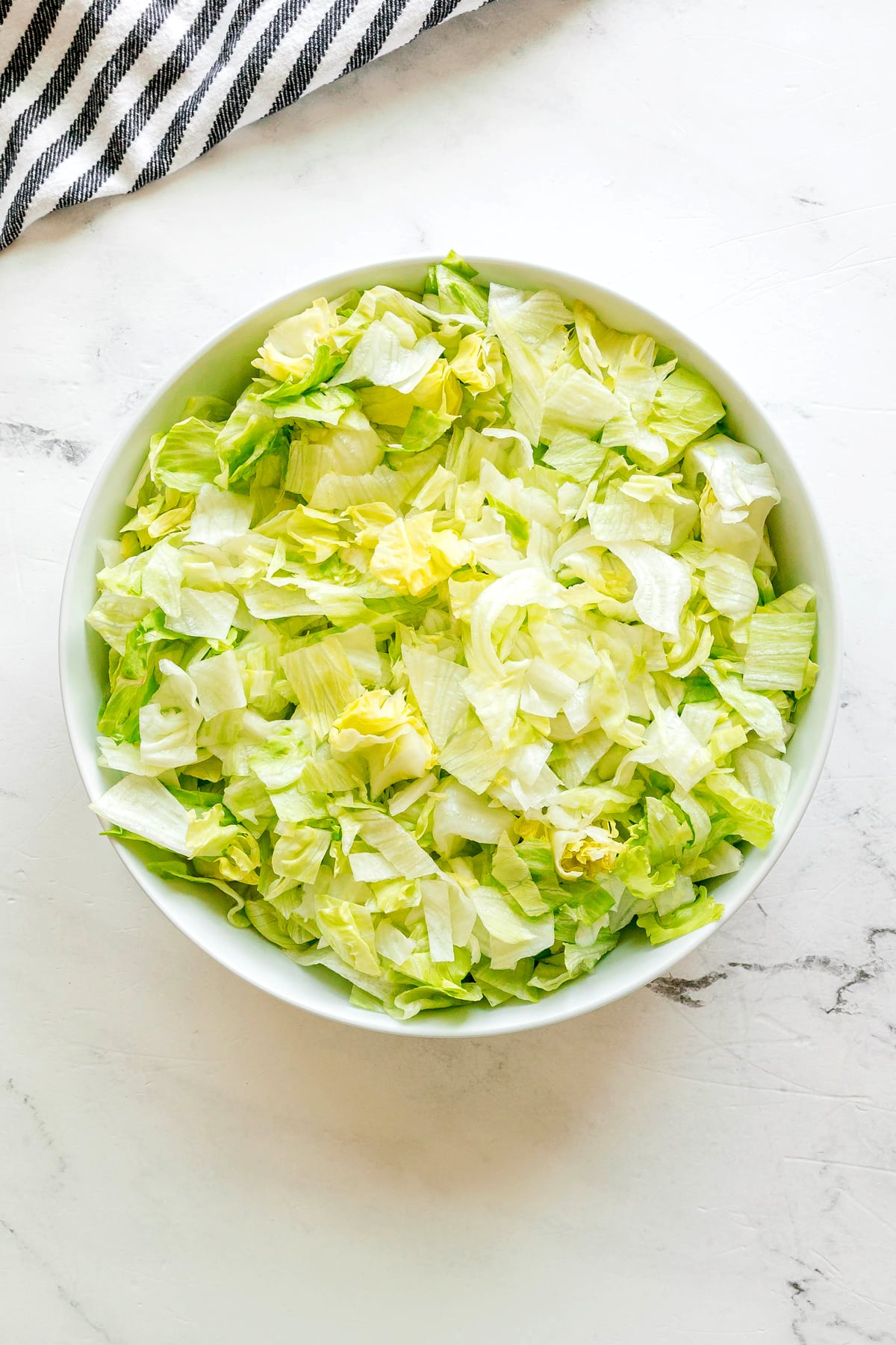 A white bowl filled with chopped iceberg lettuce for Big Mac Salad Recipe sits on a light marble surface next to a striped cloth