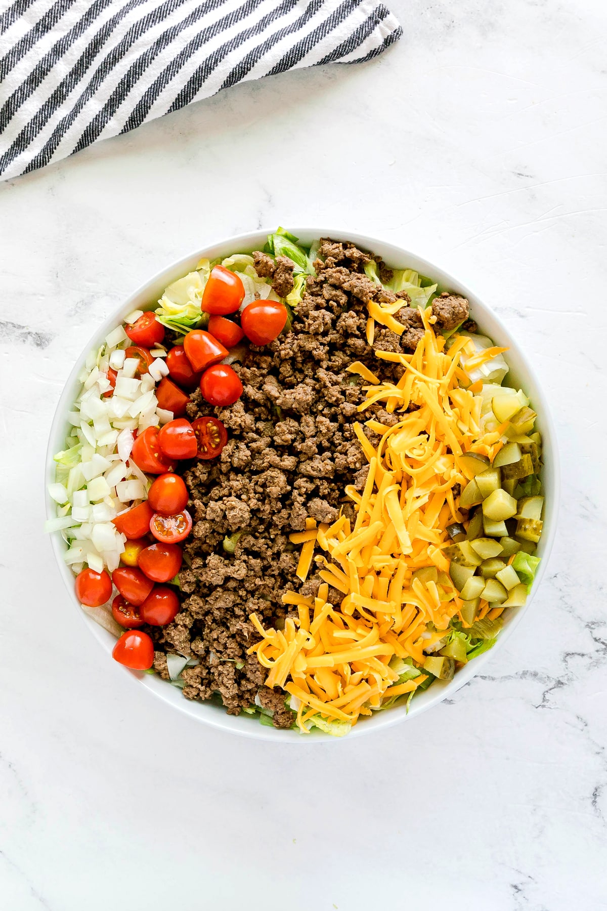 A bowl of Big Mac Salad containing sections of ground beef, shredded cheddar cheese, chopped pickles, diced onions, halved cherry tomatoes, and lettuce on a white surface.