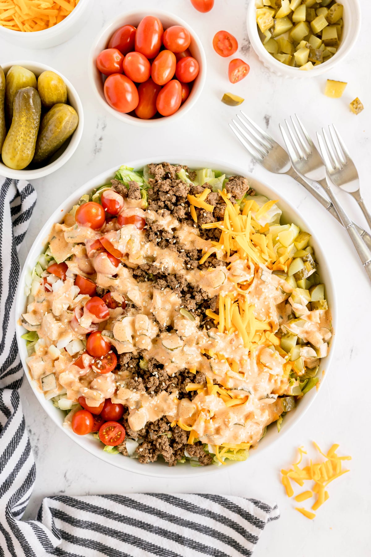 A large bowl of Big Mac Salad with ground beef, cherry tomatoes, shredded cheese, chopped pickles, and dressing, surrounded by bowls of cherry tomatoes, pickles, and forks on a white surface.