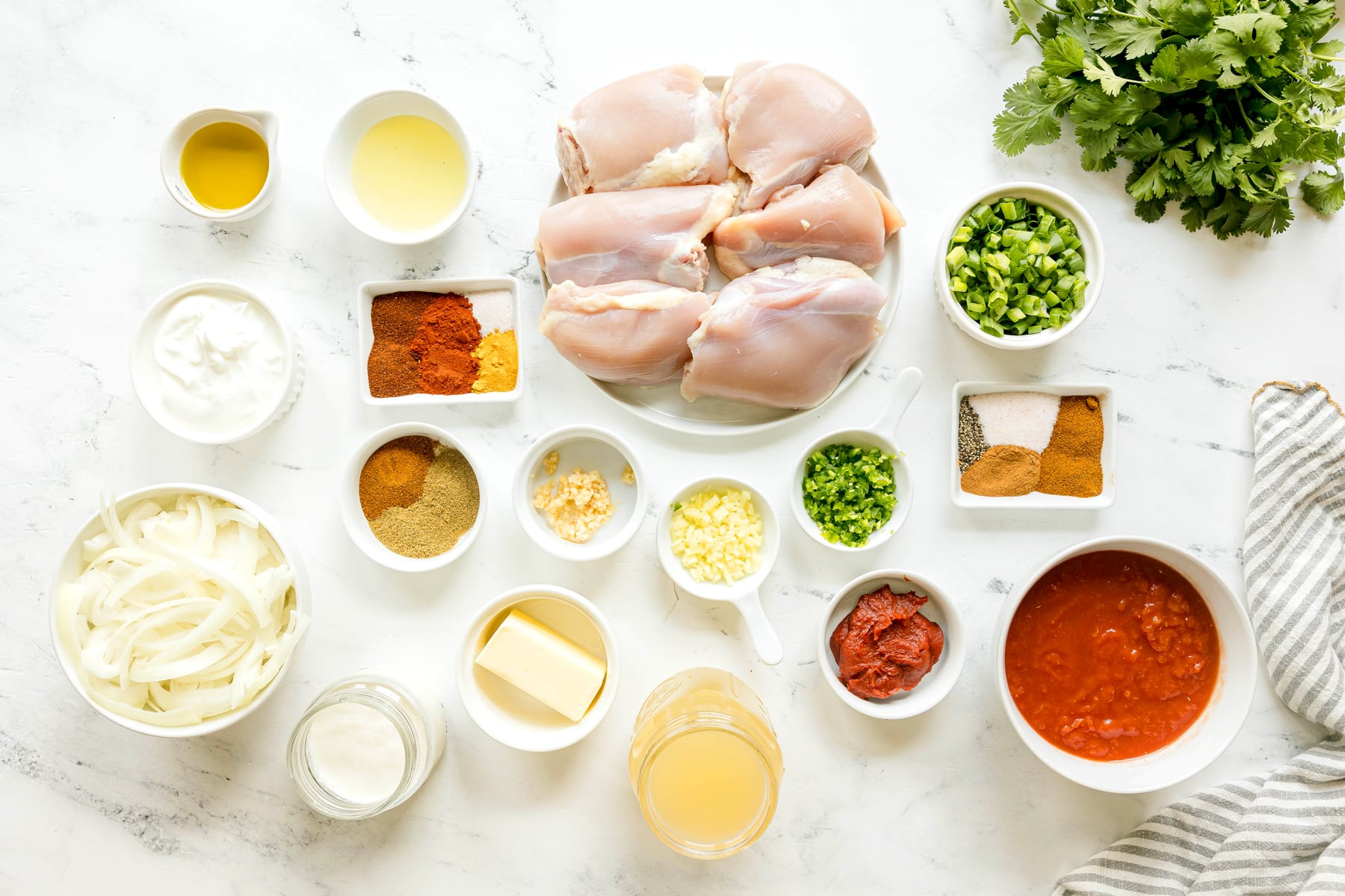 Assorted ingredients for Butter Chicken Recipe on a white surface, including olive oil, Greek yogurt, lemon juice, chili powder, paprika, salt, turmeric, garam masala, ground cumin, ground coriander, and chicken thighs, arranged in bowls and plates.