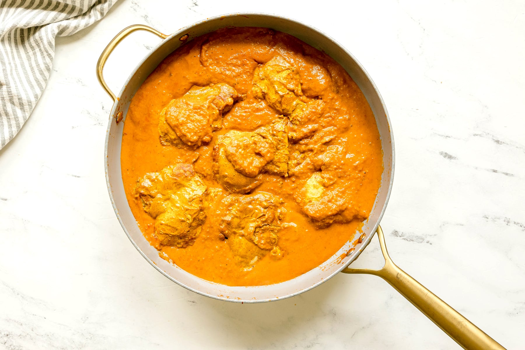 Chicken pieces simmering in a thick orange Butter Chicken sauce in a large skillet with a gold handle, set on a white marble surface.