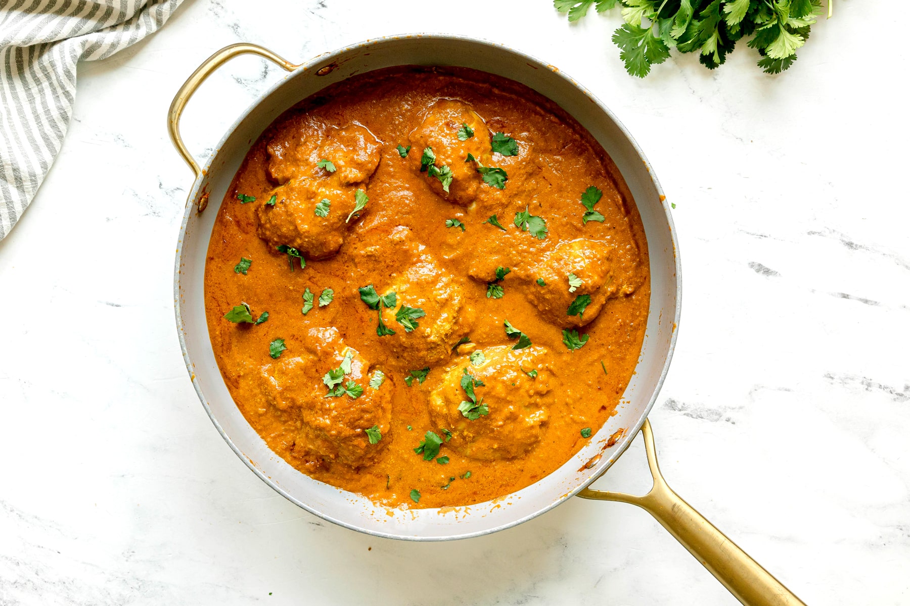 A pan filled with Butter Chicken pieces in a creamy orange sauce, garnished with chopped cilantro, sits on a marble surface.