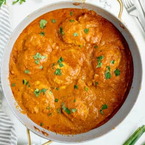 A pan filled with Butter Chicken pieces simmered in a thick, orange-brown curry sauce, garnished with chopped cilantro.